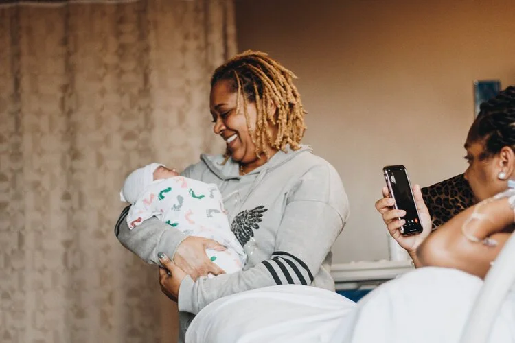 A woman holding a newborn baby wrapped in a festive blanket, smiling at the baby while another woman takes a picture with her phone in a cozy indoor setting.