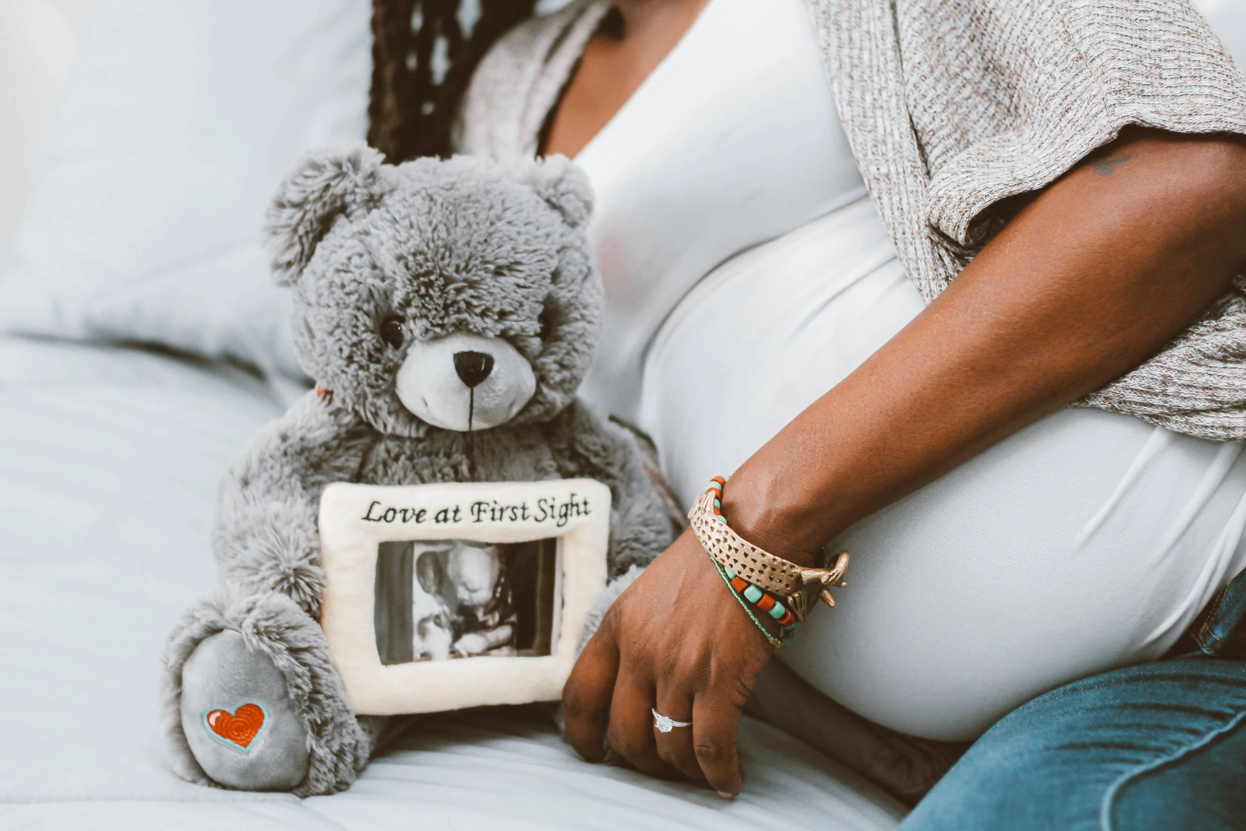 A woman sitting on a bed holding a gray teddy bear and a photo frame with the embroidered words "Love at First Sight". The woman is wearing a white shirt, jeans, and has bracelets and a ring on her hand. The teddy bear has a heart embroidery on one of its paws.