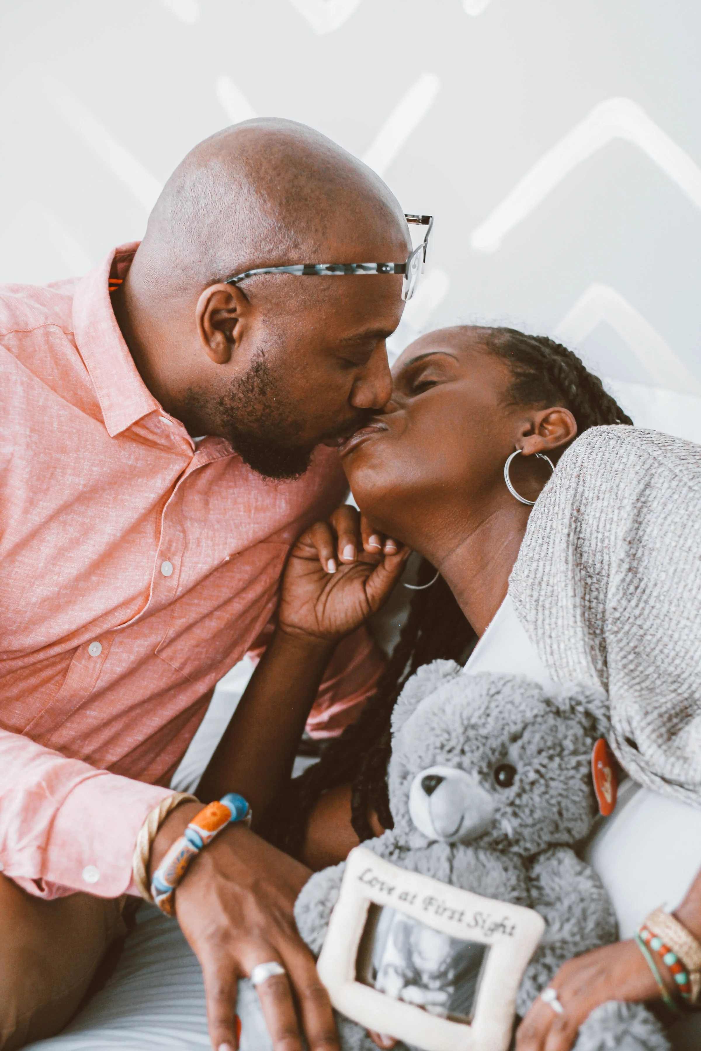 A man wearing glasses and a peach-colored shirt kisses a woman in a hospital bed, holding her hand. The woman has braided hair, hoop earrings, a gray sweater, and is holding a plush teddy bear and a framed picture with the words "Love at first sight."