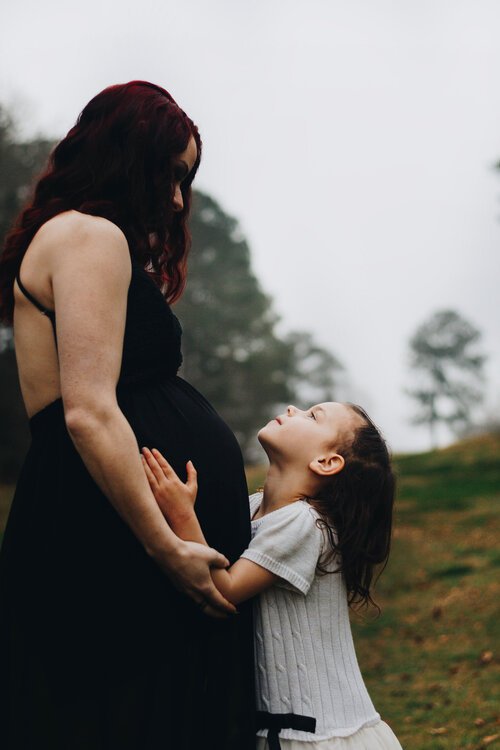 A pregnant woman and a young girl standing outdoors on a grassy area with trees in the background. The girl is looking up at the woman with admiration, and they are holding hands.