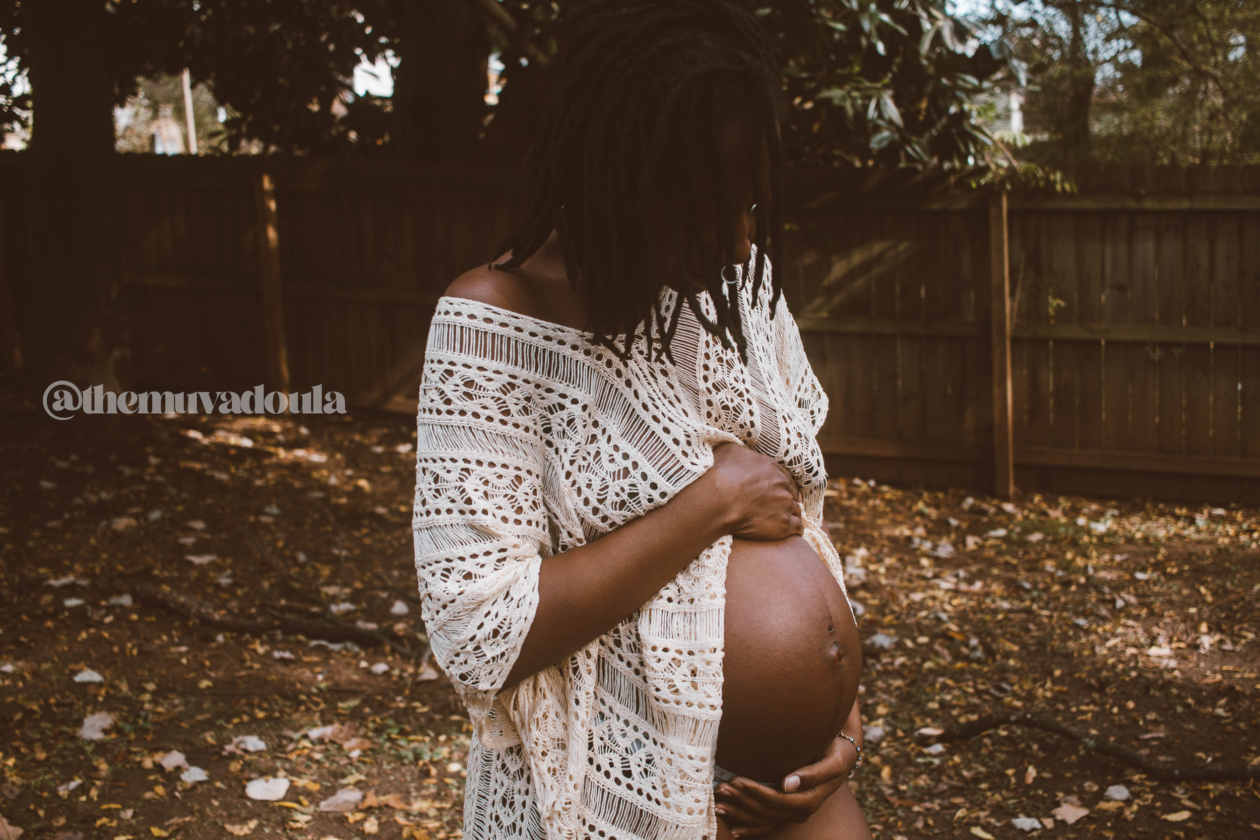 A pregnant woman wearing a lace dress standing outdoors in a backyard with a wooden fence, holding her belly and looking down.