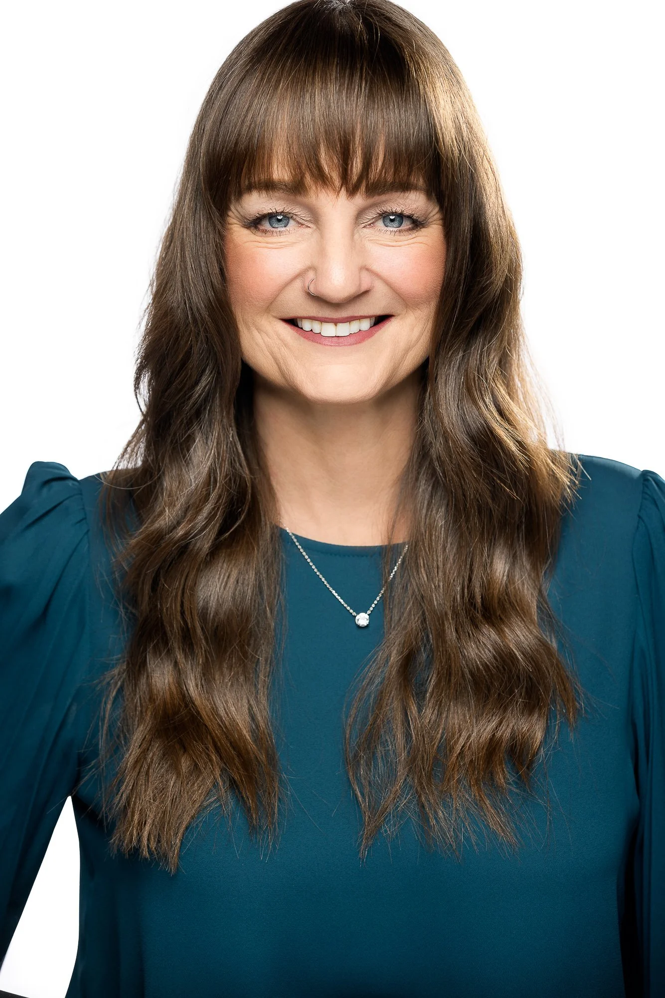 Woman with long brown hair and bangs, wearing a teal blouse and silver necklace, smiling against a white background.