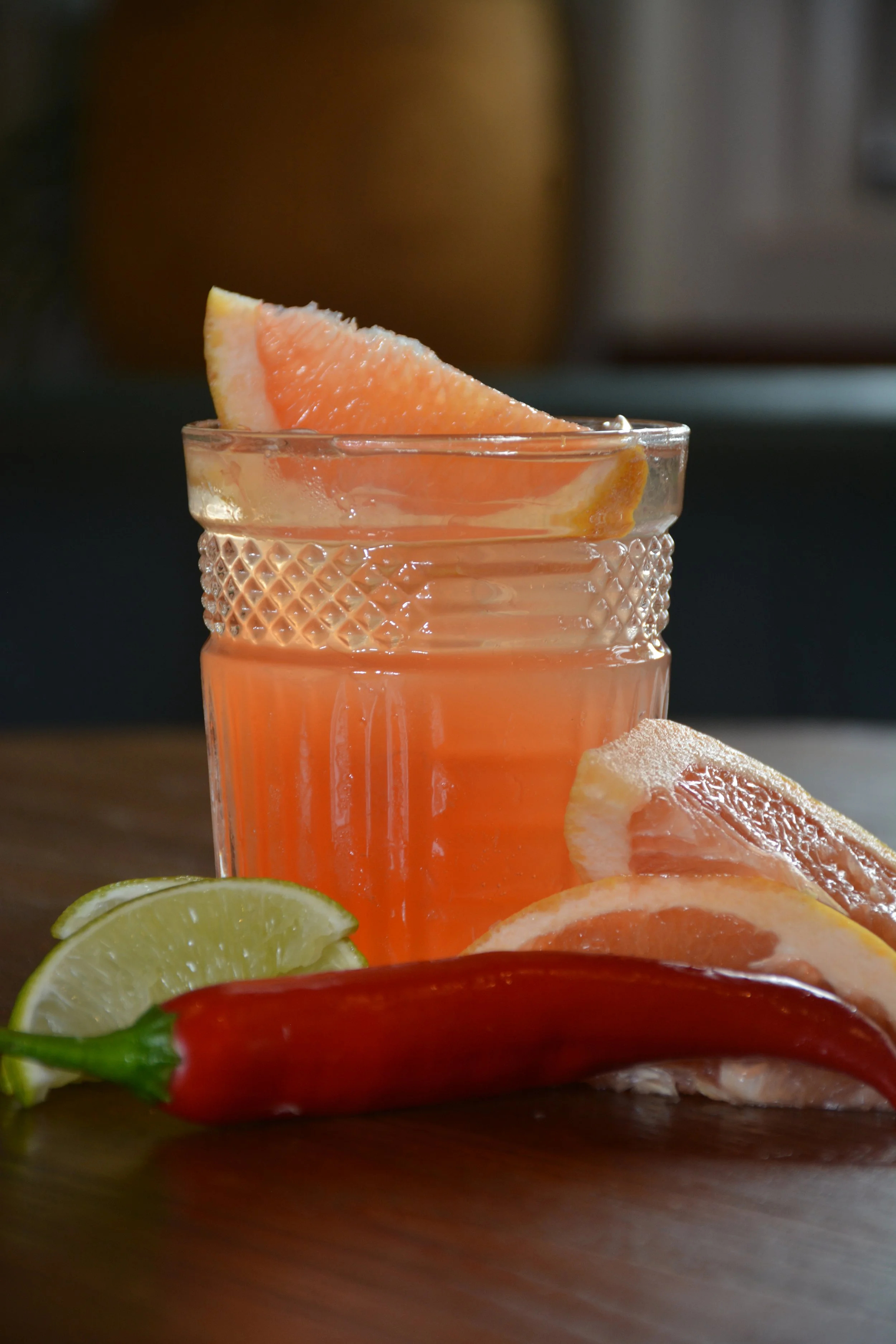 Close-up of a grapefruit slice in a textured glass, with lemon and lime slices on a wooden surface.