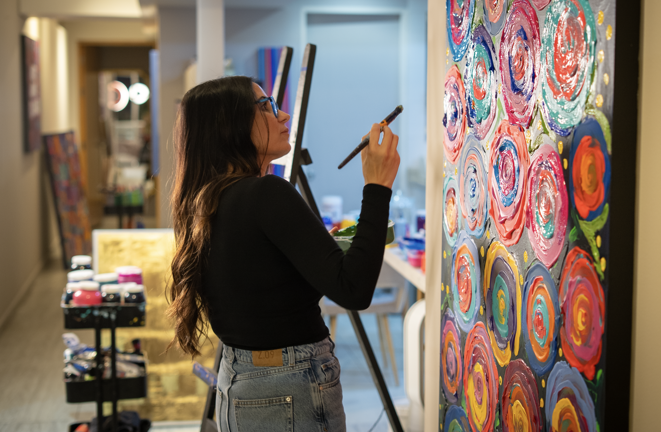 A woman with long dark hair and glasses is painting a colorful abstract floral artwork on a large canvas in an art studio.