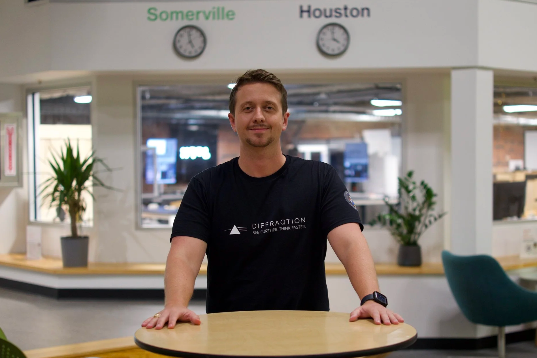 A man in a black T-shirt standing behind a round table in an office or reception area. Behind him, a sign displays the words "Somerville" and "Houston" with clocks showing different times. There are plants and office equipment visible in the background.
