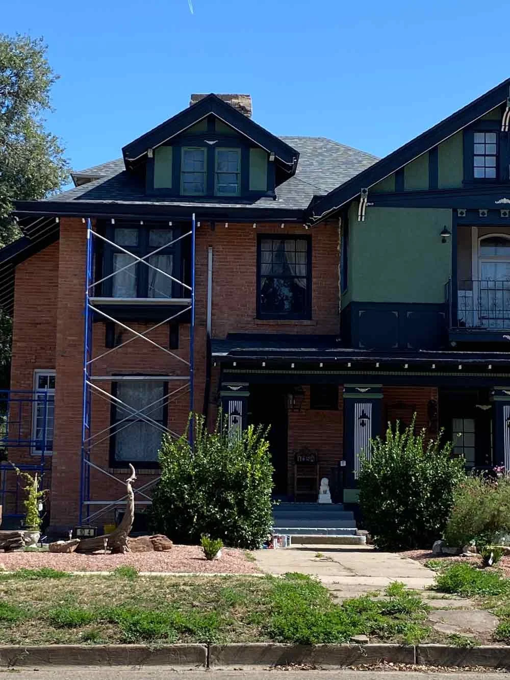 A two-story house with a brick facade on the left and a painted green and black exterior on the right. The house has a steep roof, dormer window, and a porch with steps leading up to the front door. There are bushes, plants, and a small landscaped yard in front of the house, with some construction scaffolding on the left side.
