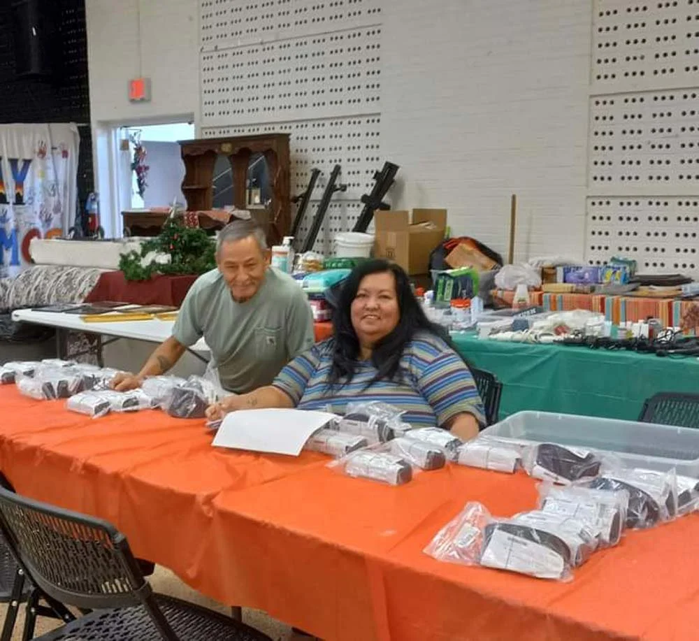Two smiling people, a man and a woman, sitting at a table covered with orange cloth, with packaged items in front of them. The background shows a room with various objects and supplies.