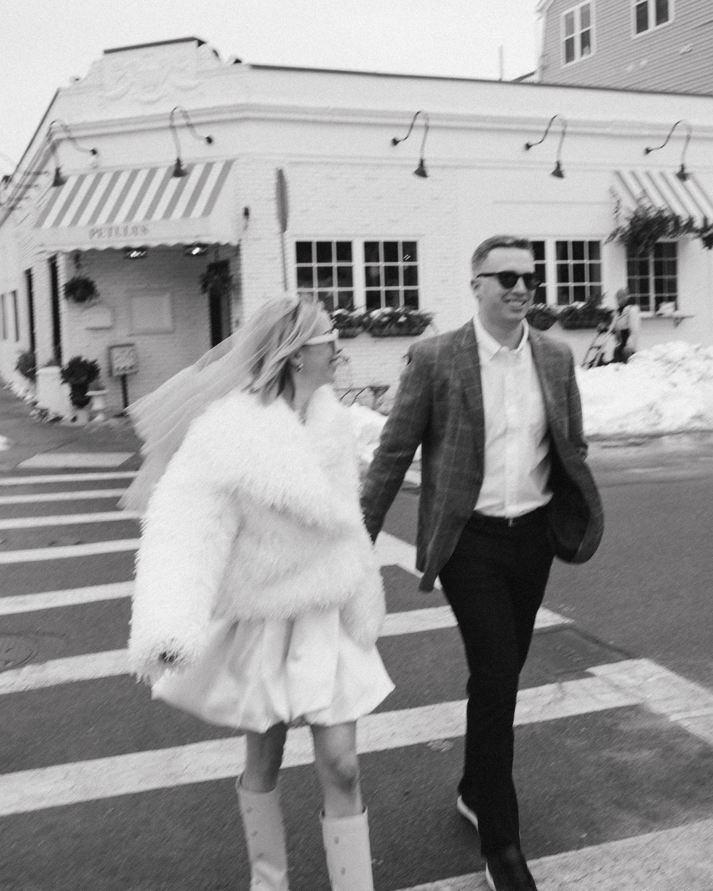 couple holds hands and crosses the crosswalk together in Boston on a snowy day - she wears a veil and boots and they are both wearing sunglasses