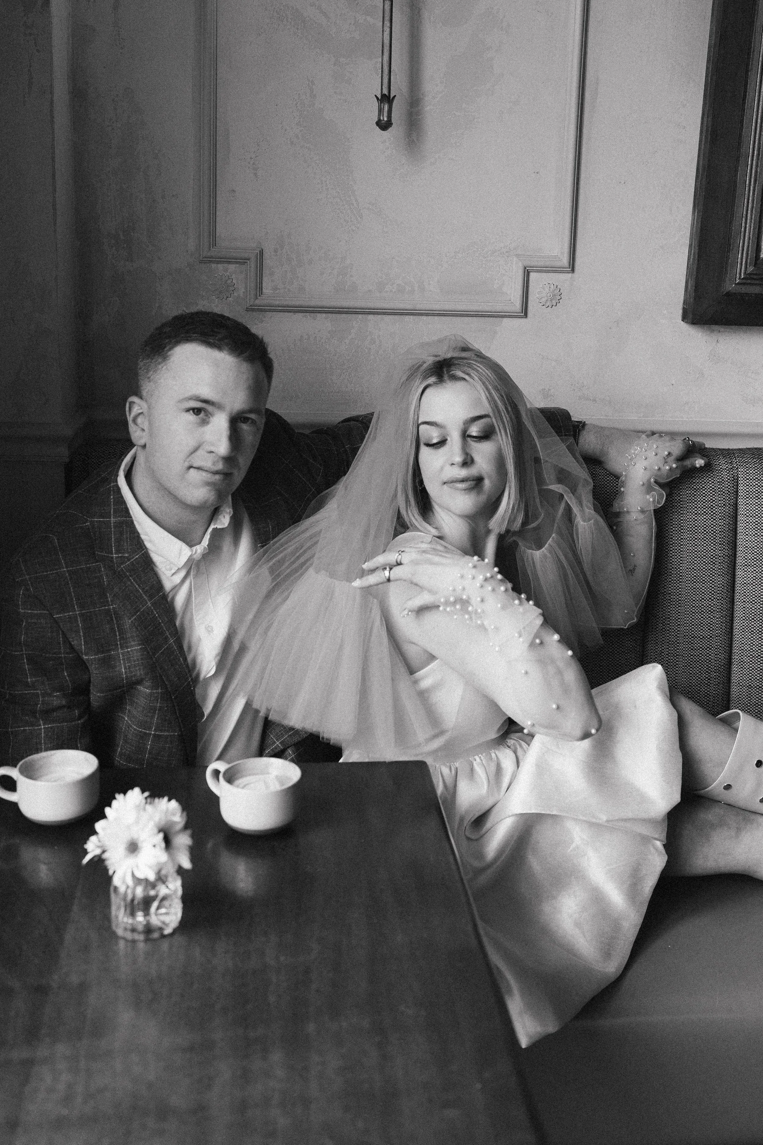woman fixing her veil while her fiance looks at the camera - they are in a coffee shop, enjoying cappuccinos and celebrating their engagement.