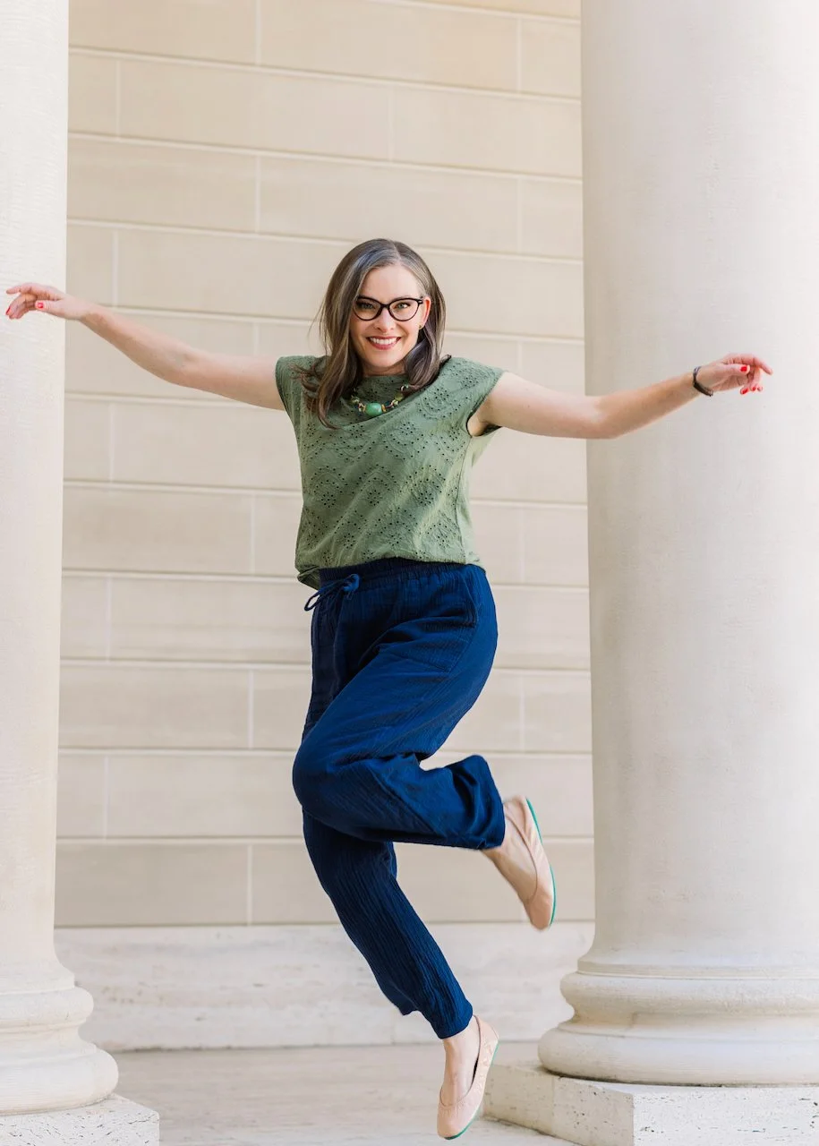 A cheerful woman with glasses, wearing a green top and blue pants, is standing between two large white columns with her arms outstretched.