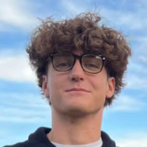 Portrait of a young man with curly hair wearing glasses, against a blue sky background.