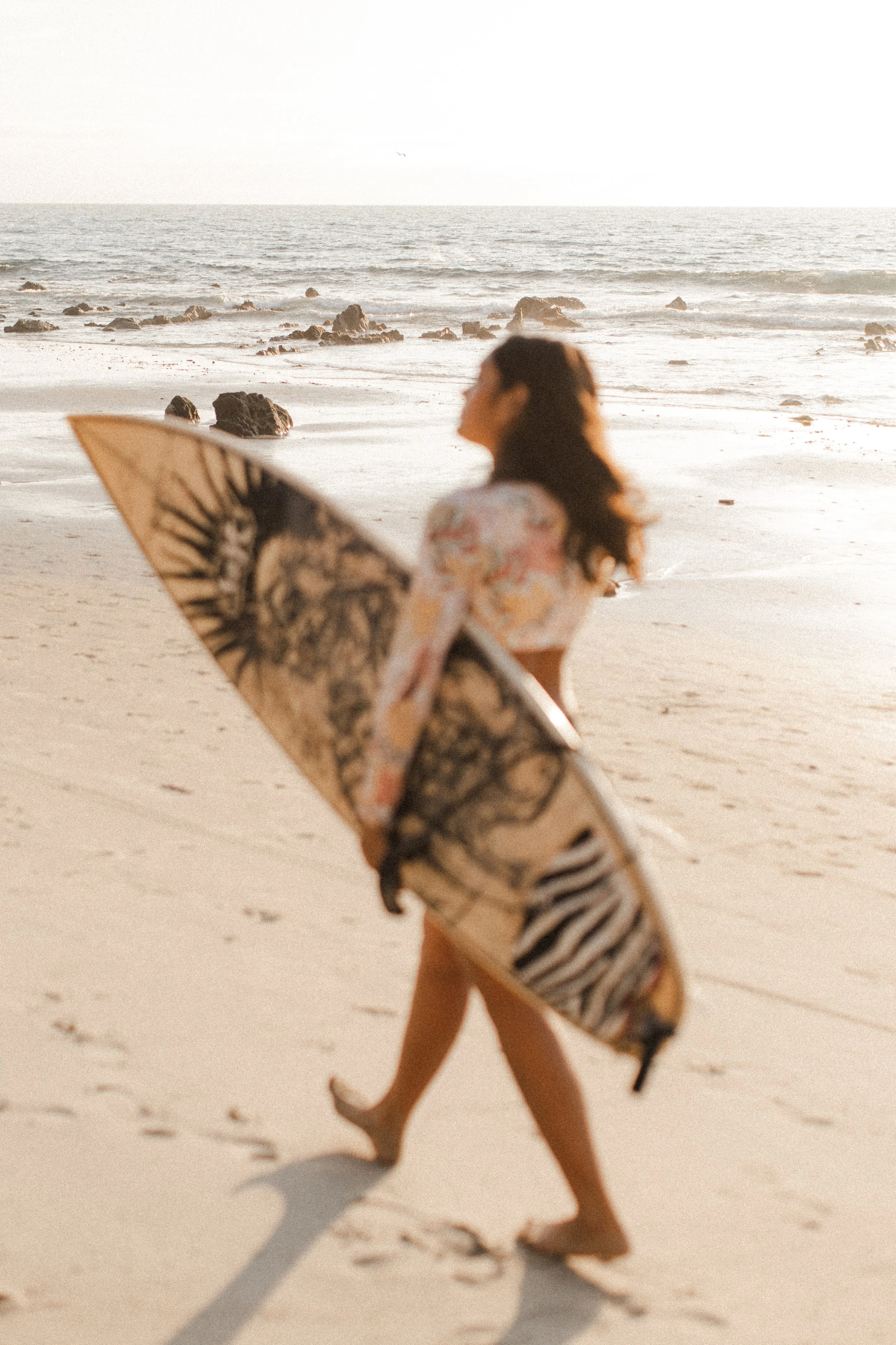 A woman walking on the beach holding a surfboard, with the ocean and rocks in the background, during sunset.
