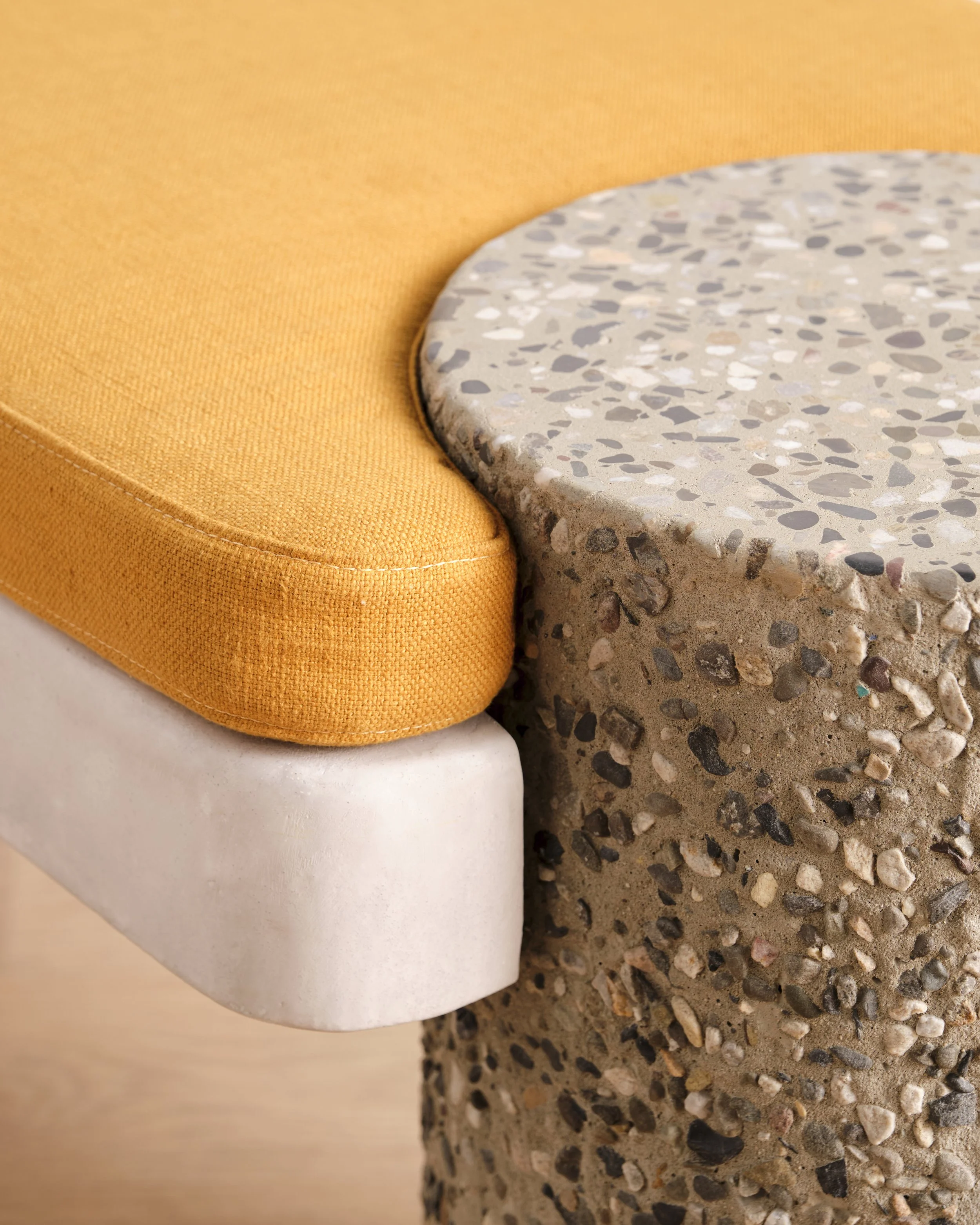 Close-up detail of the Betty Bench, a modern bench with an ochre-colored cushion, hand-sculpted base, and a pebble-textured concrete column leg on a light wooden floor against a plain white wall.