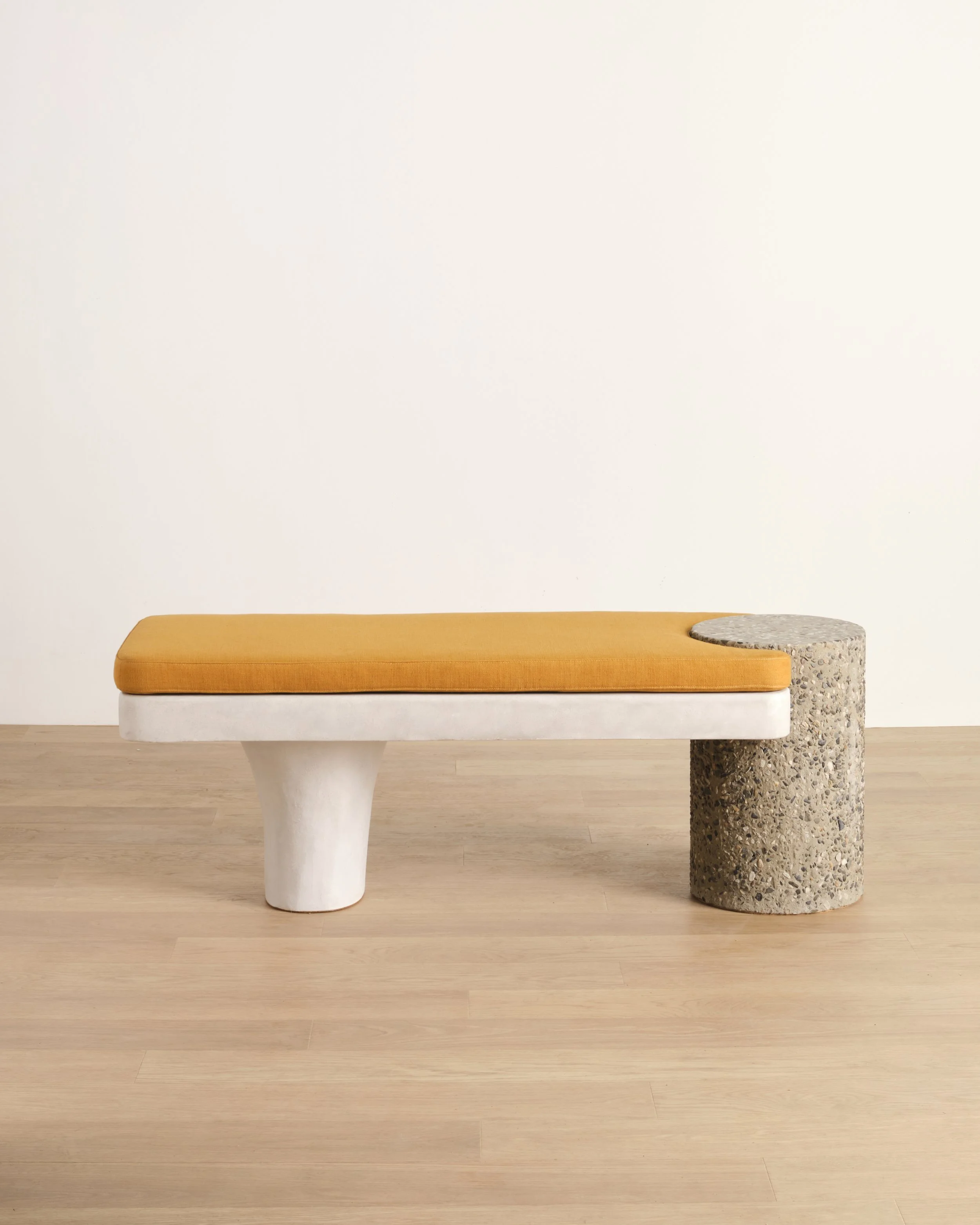 Betty Bench, a modern bench with an ochre-colored cushion, hand-sculpted base, and a pebble-textured concrete column leg on a light wooden floor against a plain white wall.