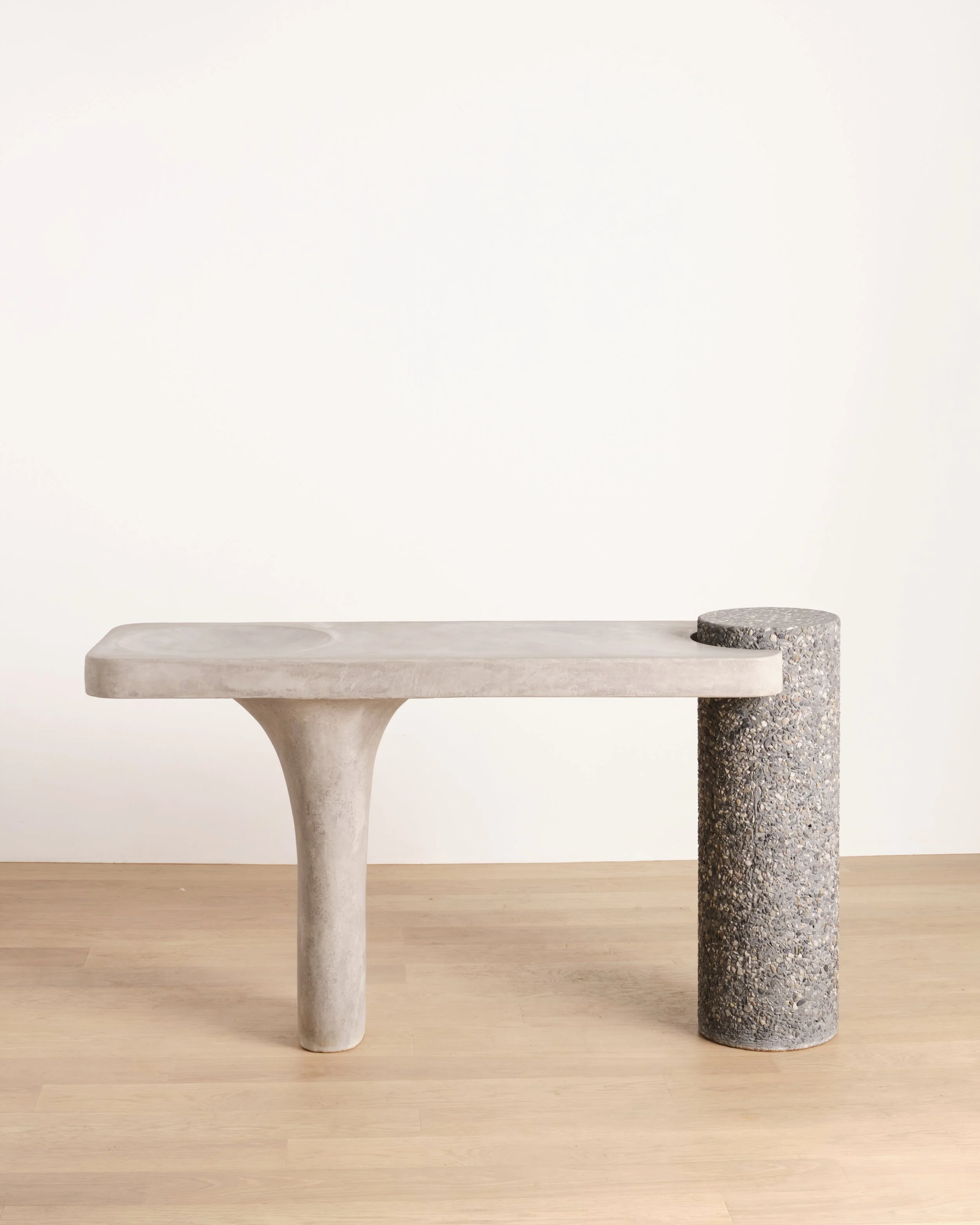 A minimalistic bench with a stone-like surface and a cylindrical stone support on a light wooden floor against a plain white wall.