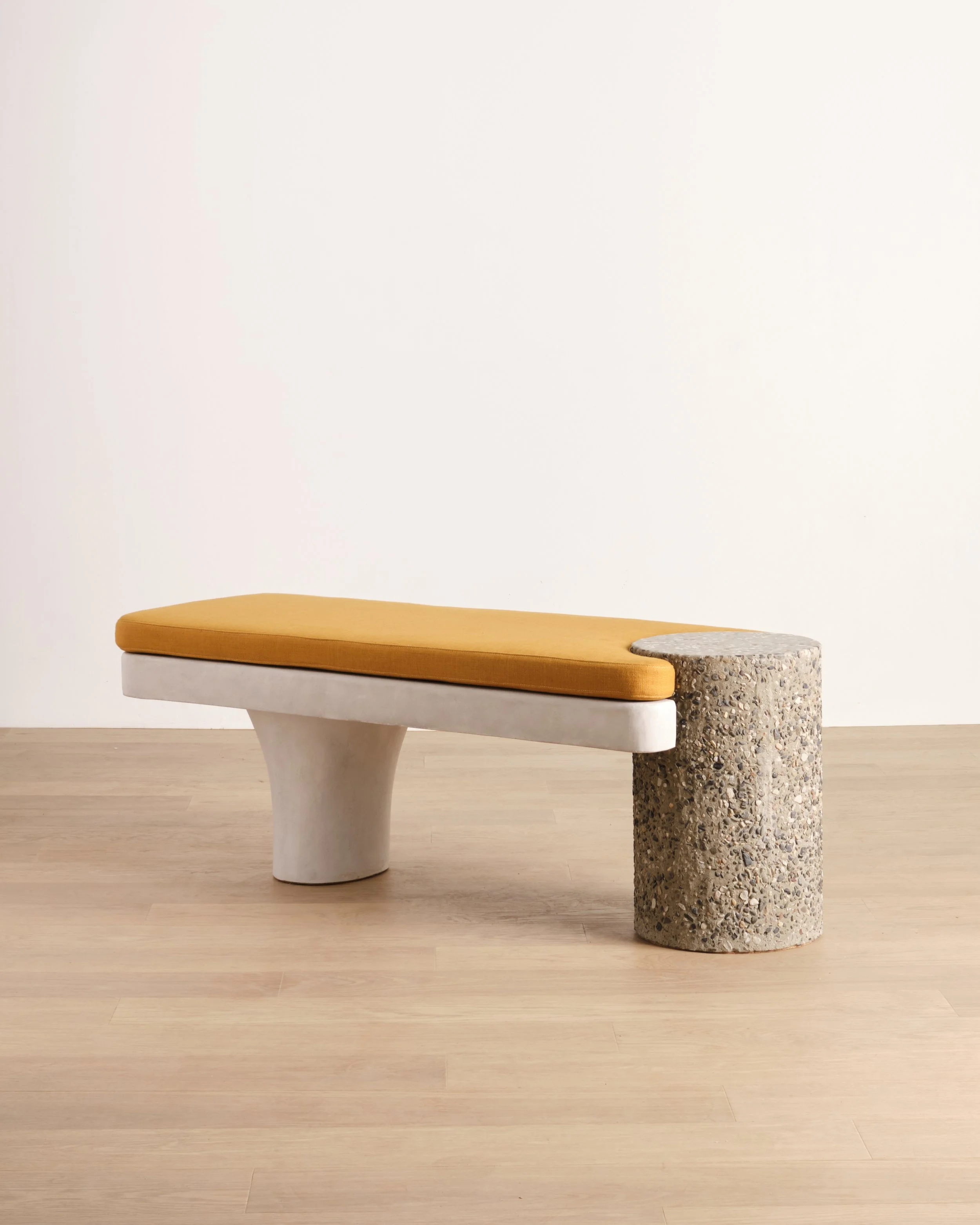 Betty Bench, a modern bench with an ochre-colored cushion, hand-sculpted base, and a pebble-textured concrete column leg on a light wooden floor against a plain white wall.