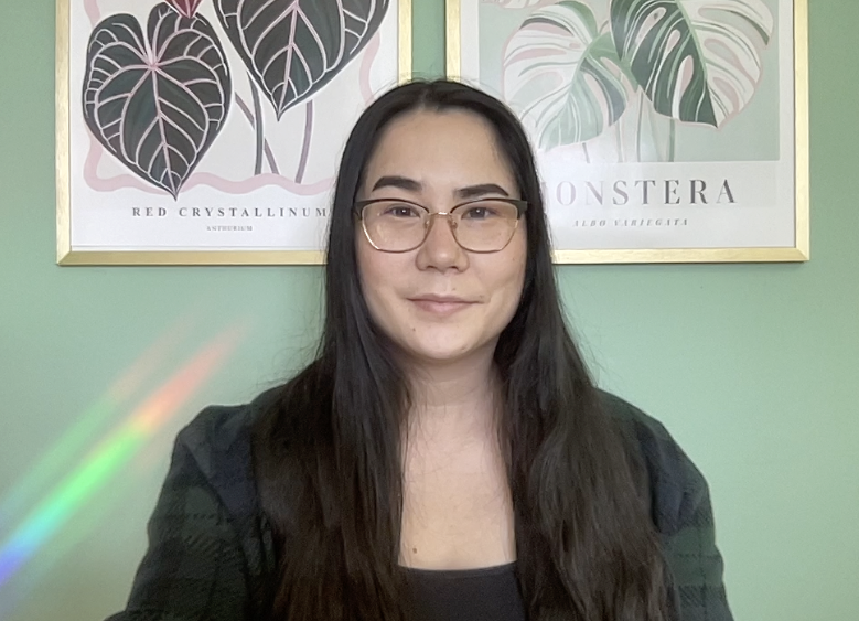A woman with long dark hair and glasses smiling at the camera, with two framed botanical prints of tropical leaves hanging on a light green wall behind her.