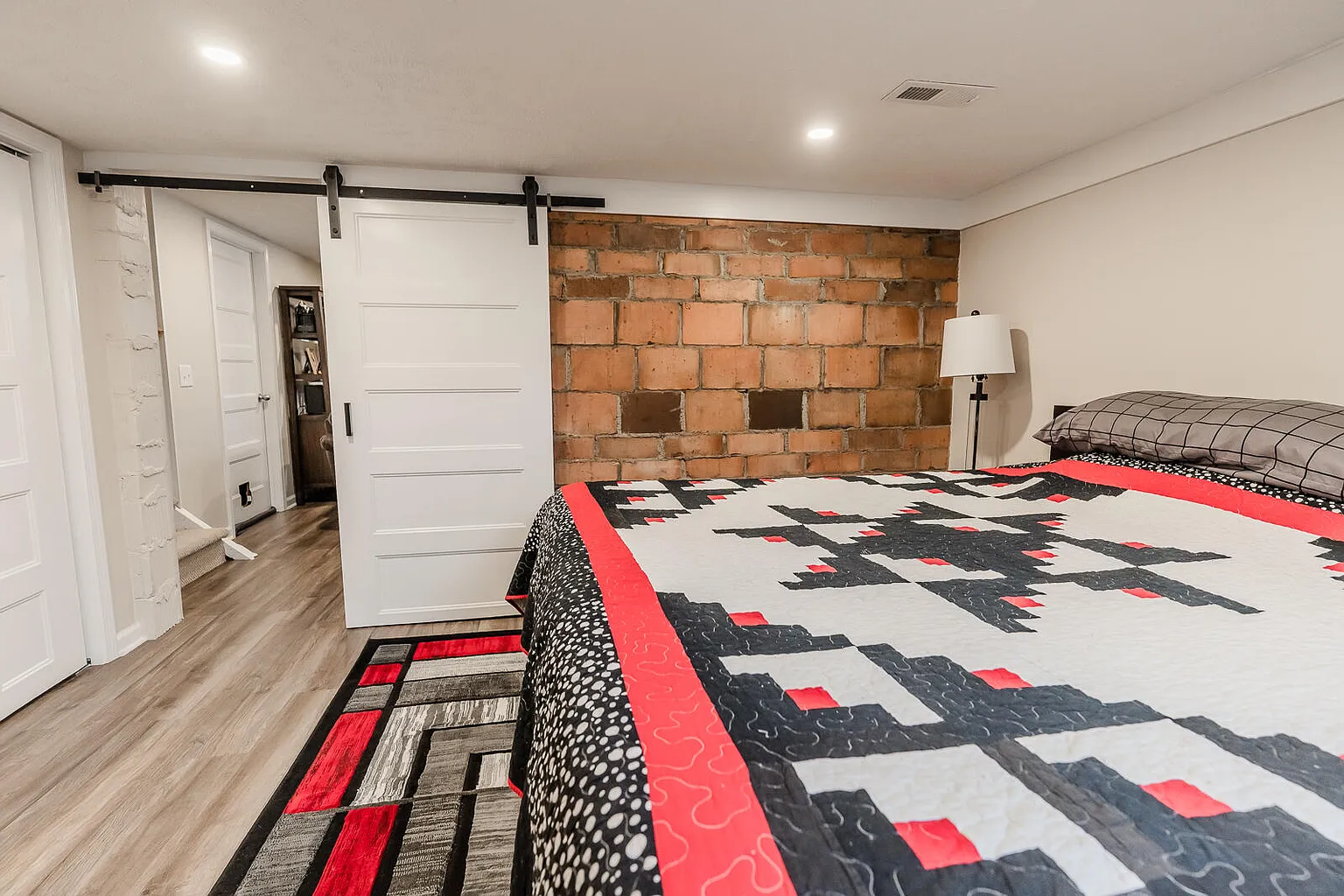 A bedroom with a large bed covered in a quilt with black, white, and red geometric patterns, a brick accent wall, a white sliding barn door, and a floor lamp beside the bed.