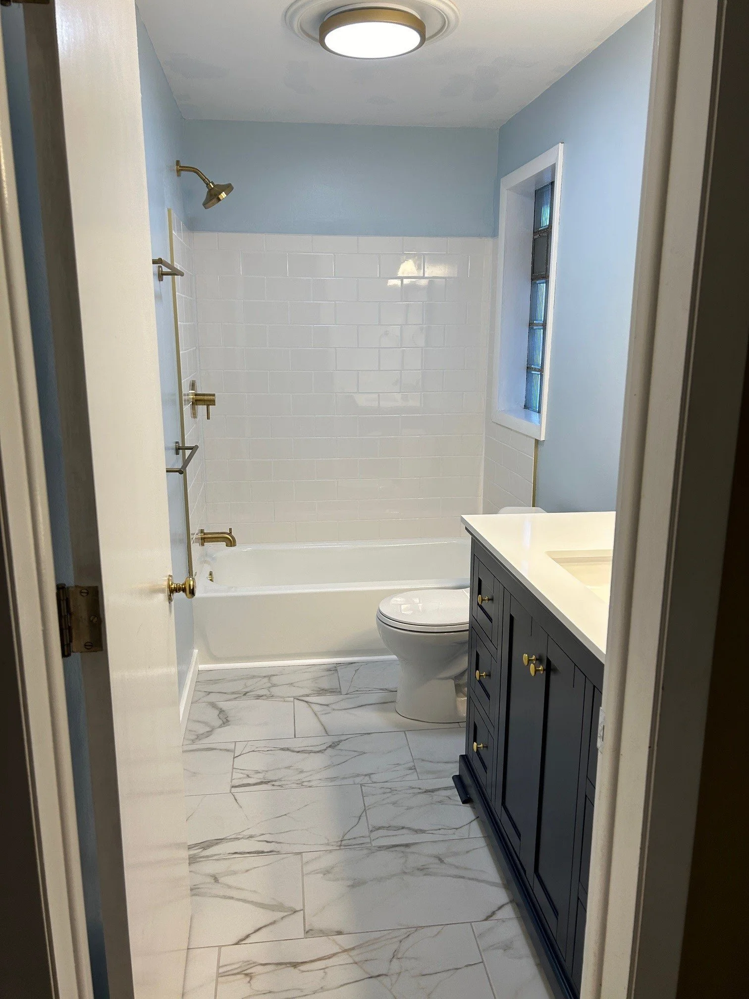 A modern bathroom featuring a walk-in bathtub with tile walls, a toilet, a vanity with gold knobs, and marble-look floor tiles, illuminated by a ceiling light.