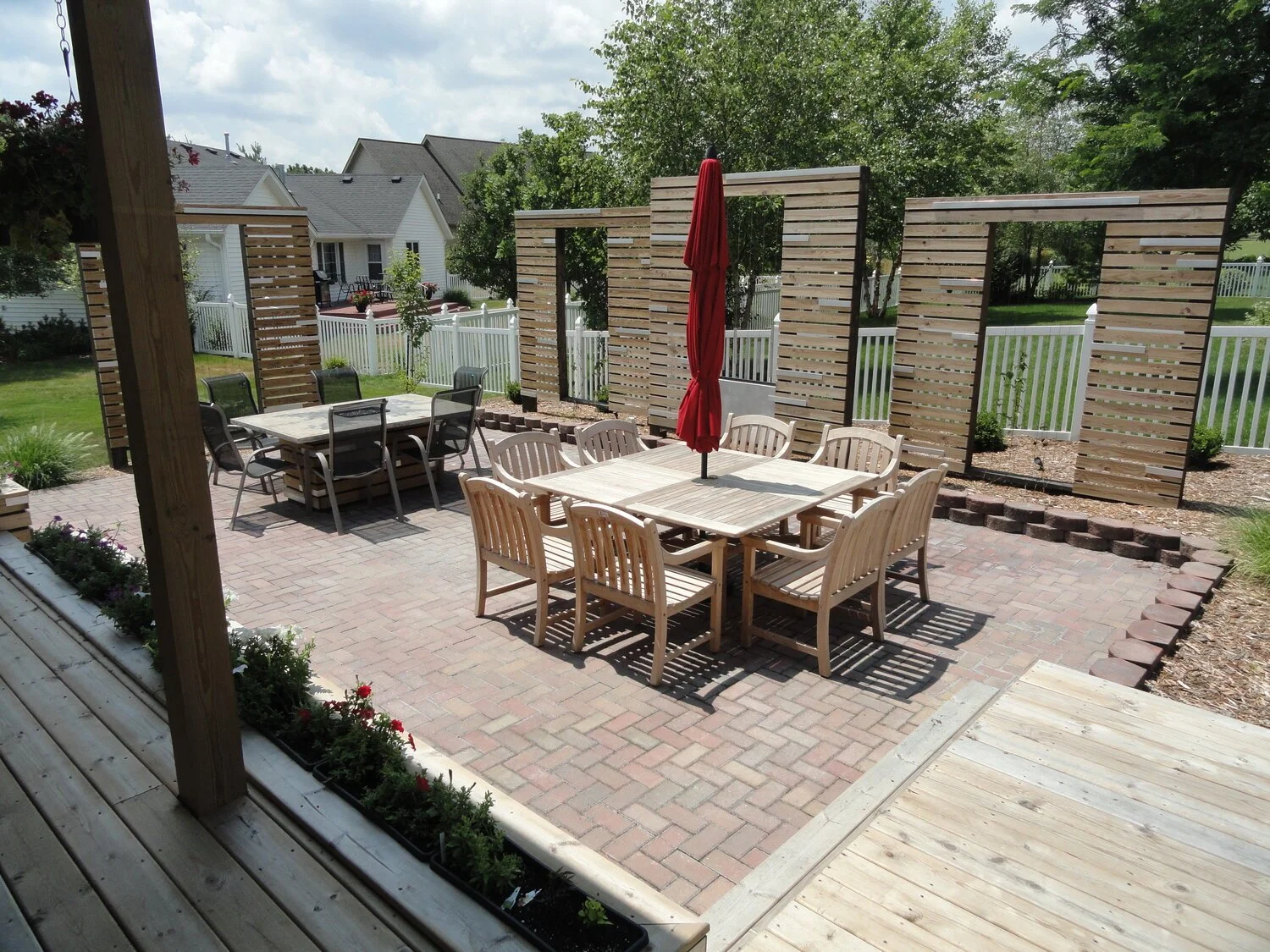 A backyard patio with brick flooring, wooden dining table with chairs, and a closed red umbrella. Wooden privacy screens and a white fence surround the yard, with trees and neighboring houses in the background.