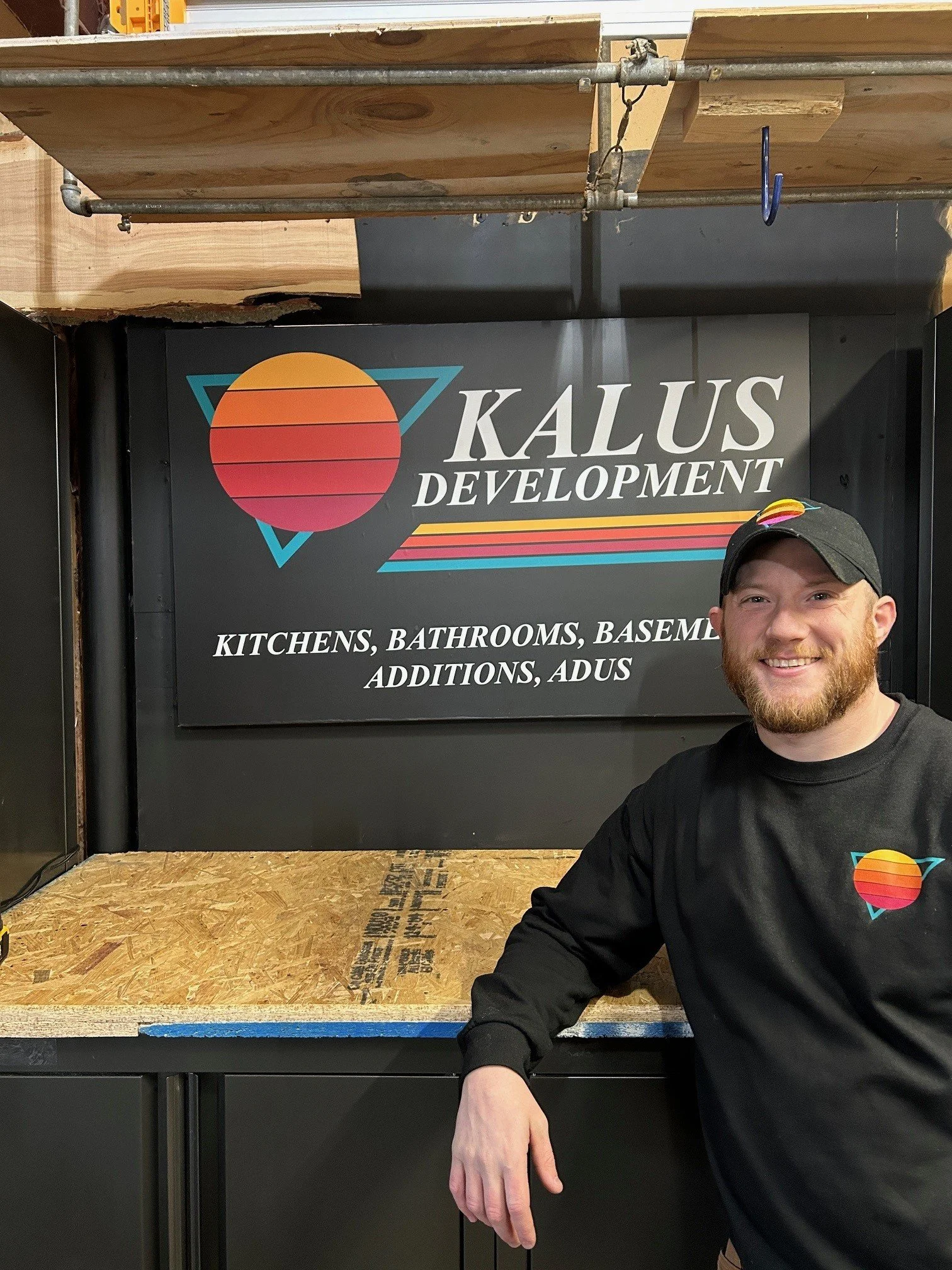 Man with a beard and black cap smiling, standing next to a black sign with a colorful logo, that reads 'KALUS DEVELOPMENT' and lists services like kitchens, bathrooms, basements, additions, and ADUs.