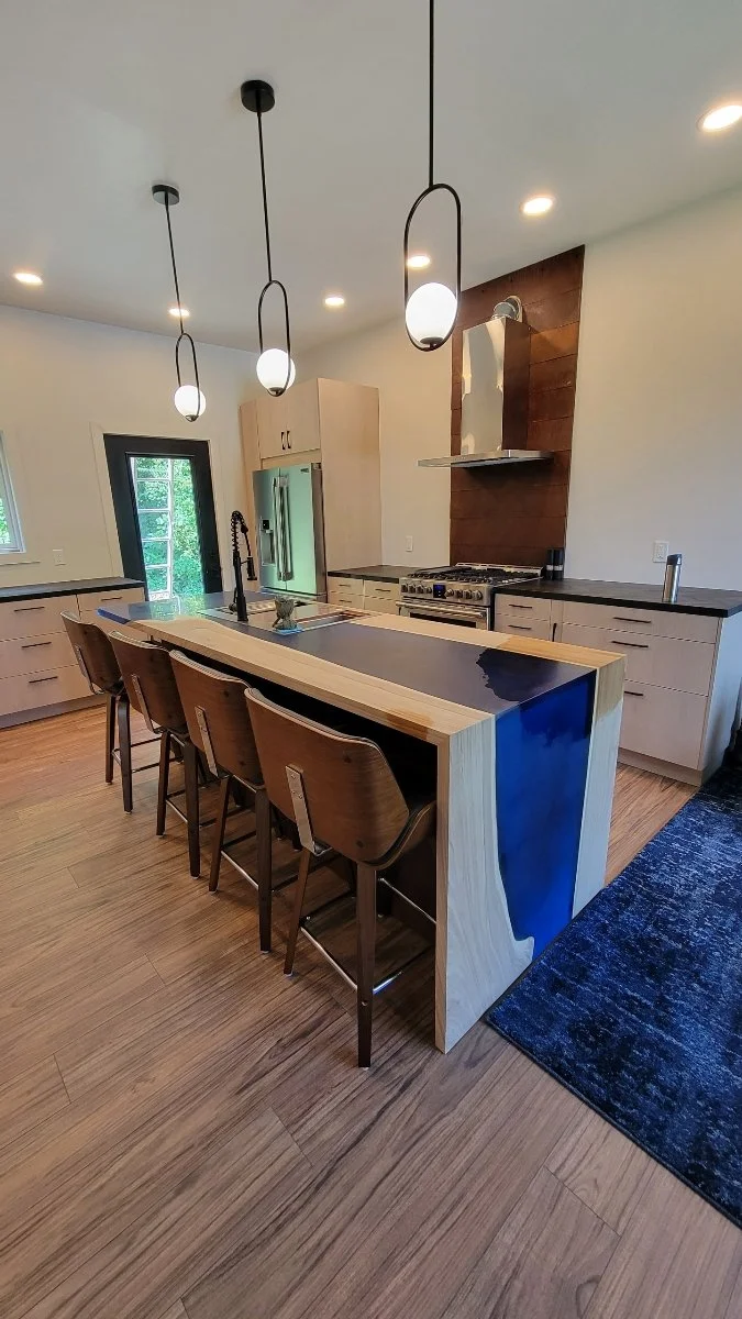 Modern kitchen with a wooden island featuring a black countertop, four bar stools, pendant lighting, a stainless steel refrigerator, a stove with a wooden backsplash, and a black rug on a wood floor.