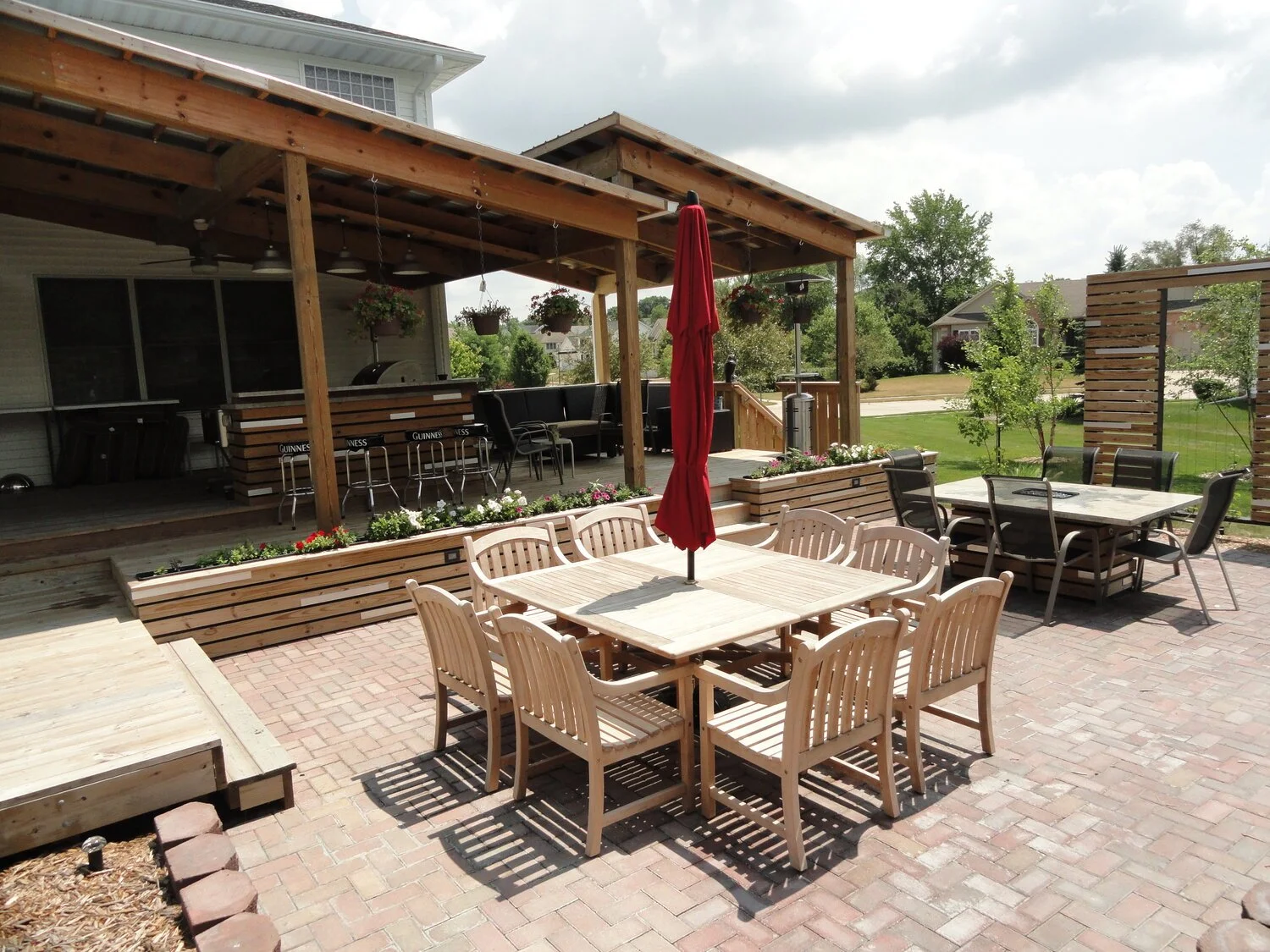 Outdoor backyard patio with wooden tables and chairs, a patio umbrella, and a covered deck with hanging flower baskets and bar seating.
