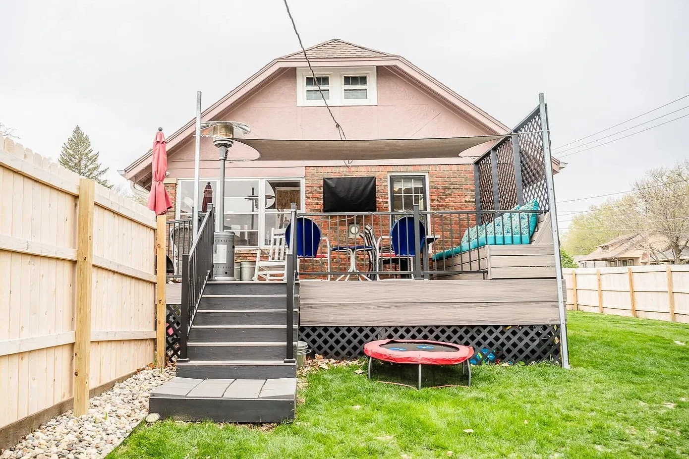 Backyard with wooden deck, fenced yard, chairs, a trampoline, and a house with a brick and siding exterior.