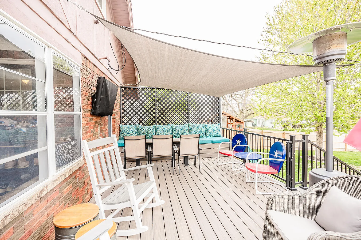 Outdoor patio with seating area, including chairs, a wicker lounge chair with pillow, a bench with colorful cushions, and children's chairs with star decoration, covered by a beige shade sail, with a brick exterior wall and trees in the background.