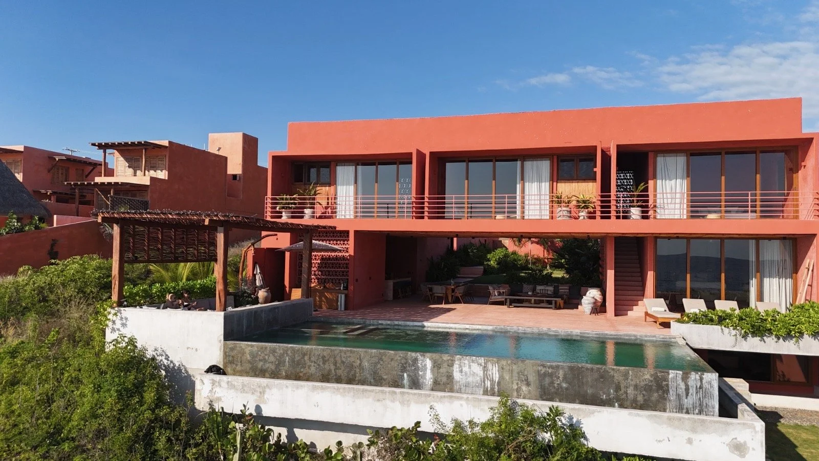 Modern multi-story house with large glass windows, red and pink exterior walls, overlooking a swimming pool, with outdoor seating and lush greenery.