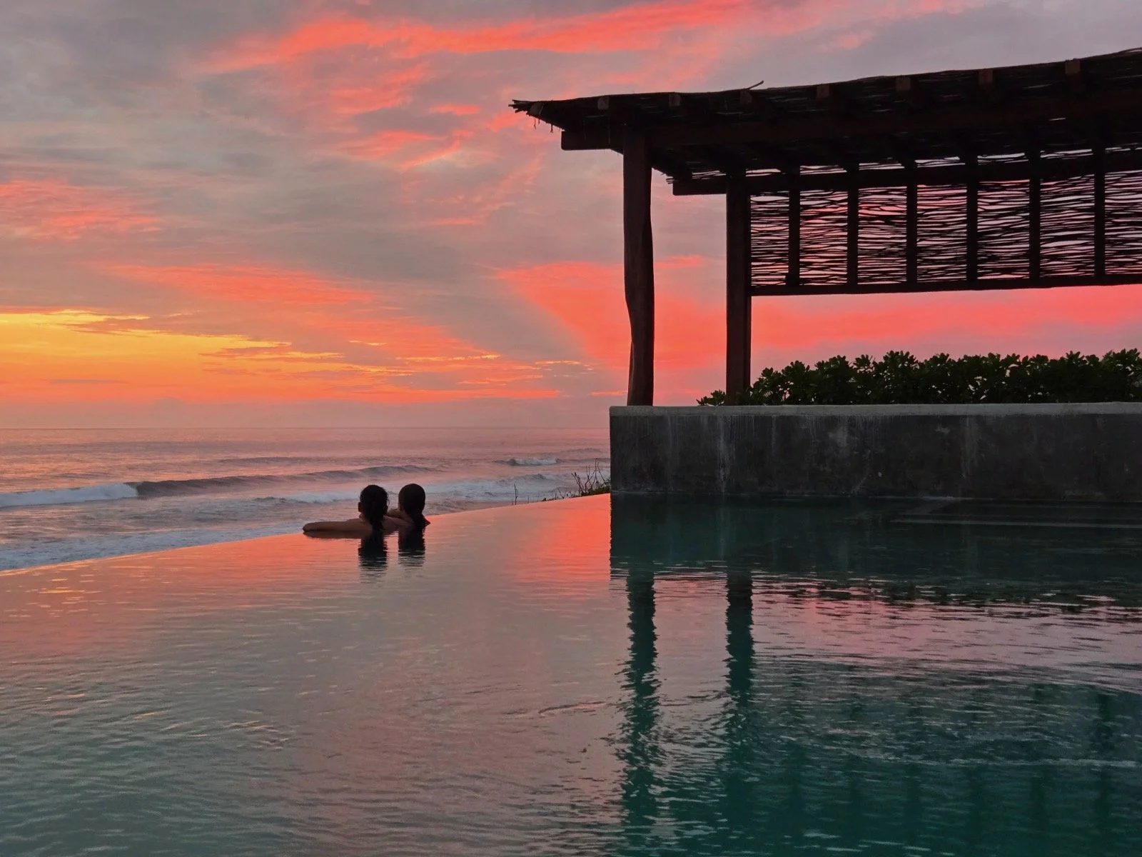 Two people are in a pool, watching the sunset by the ocean, with a thatched-roof structure nearby and pink and orange clouds in the sky.