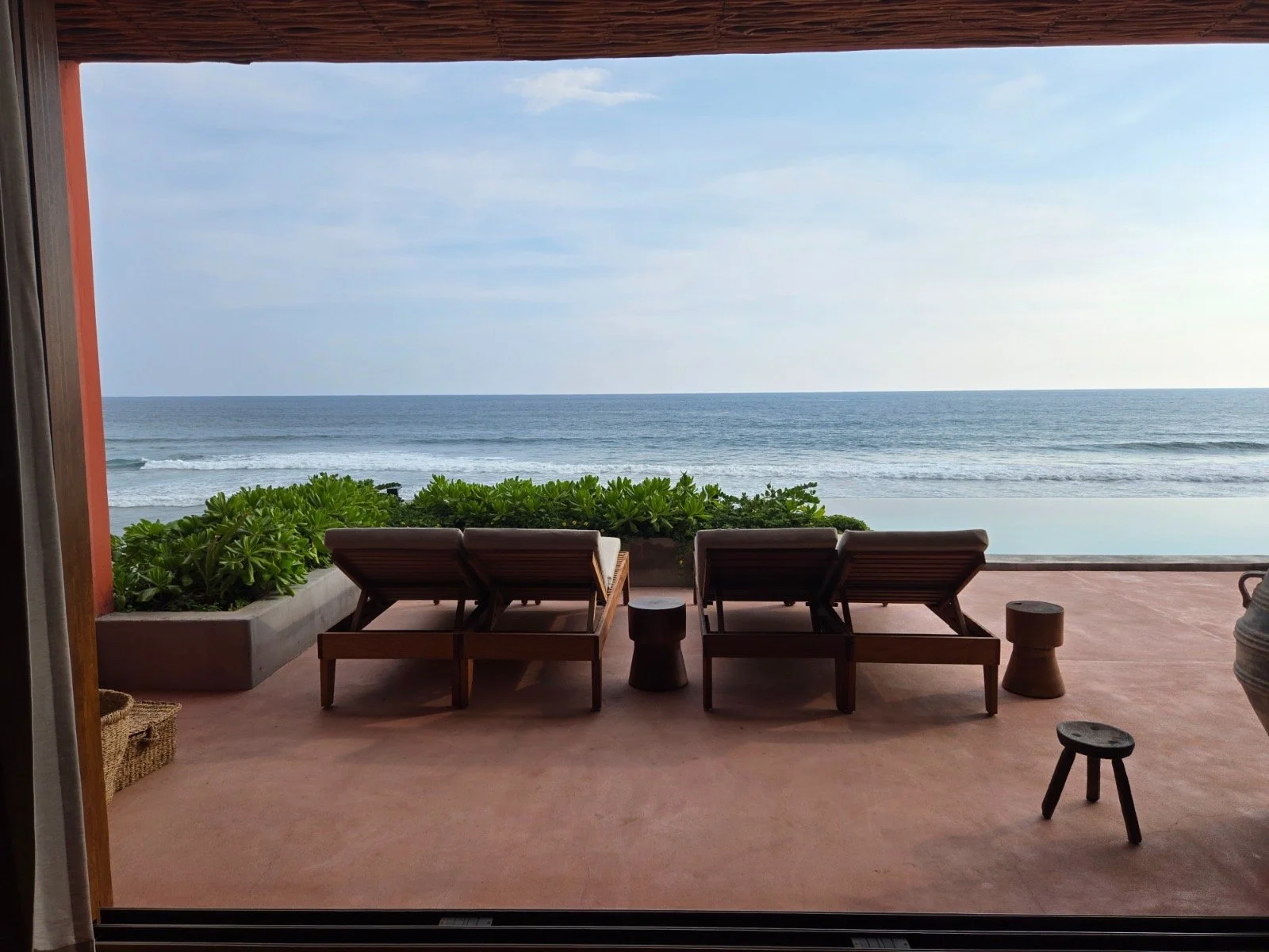 View from a patio or balcony overlooking the beach with three wooden lounge chairs, small side tables, and green plants in the foreground, ocean and sky in the background.