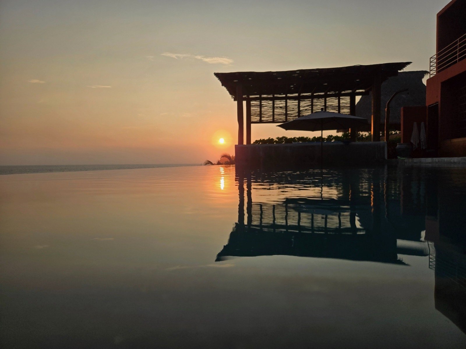 Sunset over a calm ocean with a silhouette of a structure near the water and its reflection visible in the water.