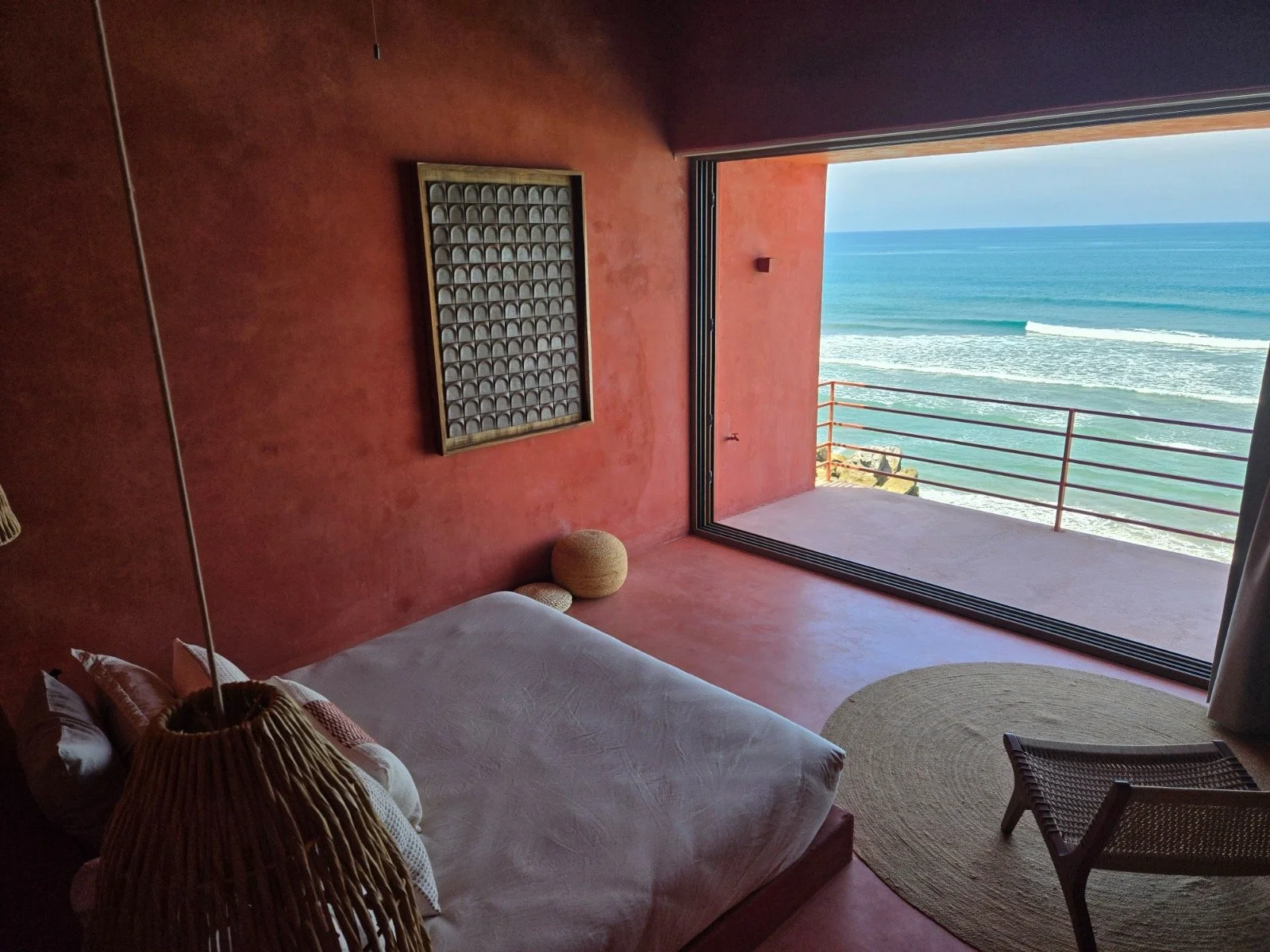 A bedroom with a large window opening to a view of the ocean and sandy beach, with a bed, wicker lamp, and woven cushions.