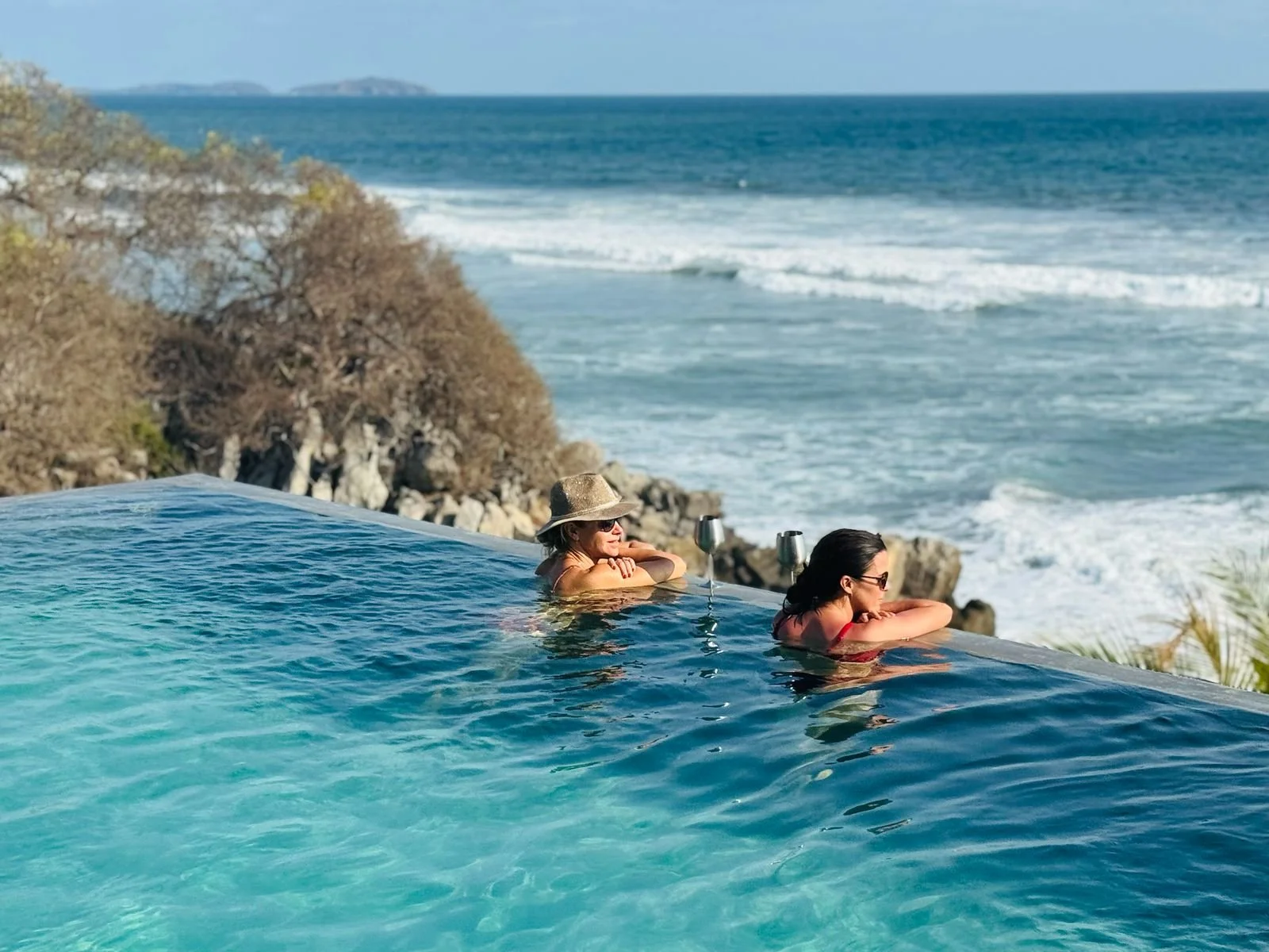 Two women relax in an infinity pool overlooking the ocean with a rocky coastline and distant islands in the background.
