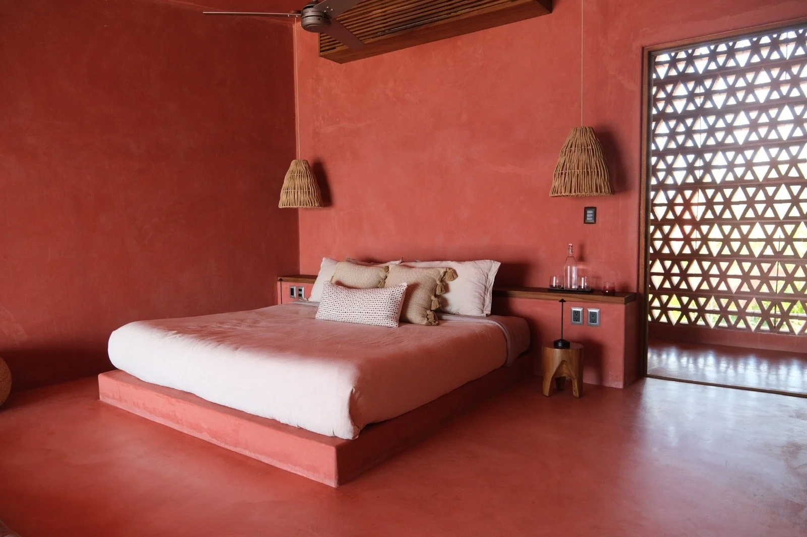A bedroom with pink walls and floor. It has a bed with white and beige pillows, wooden bedside tables, and woven pendant lights. There's a lattice window or door on the right side.