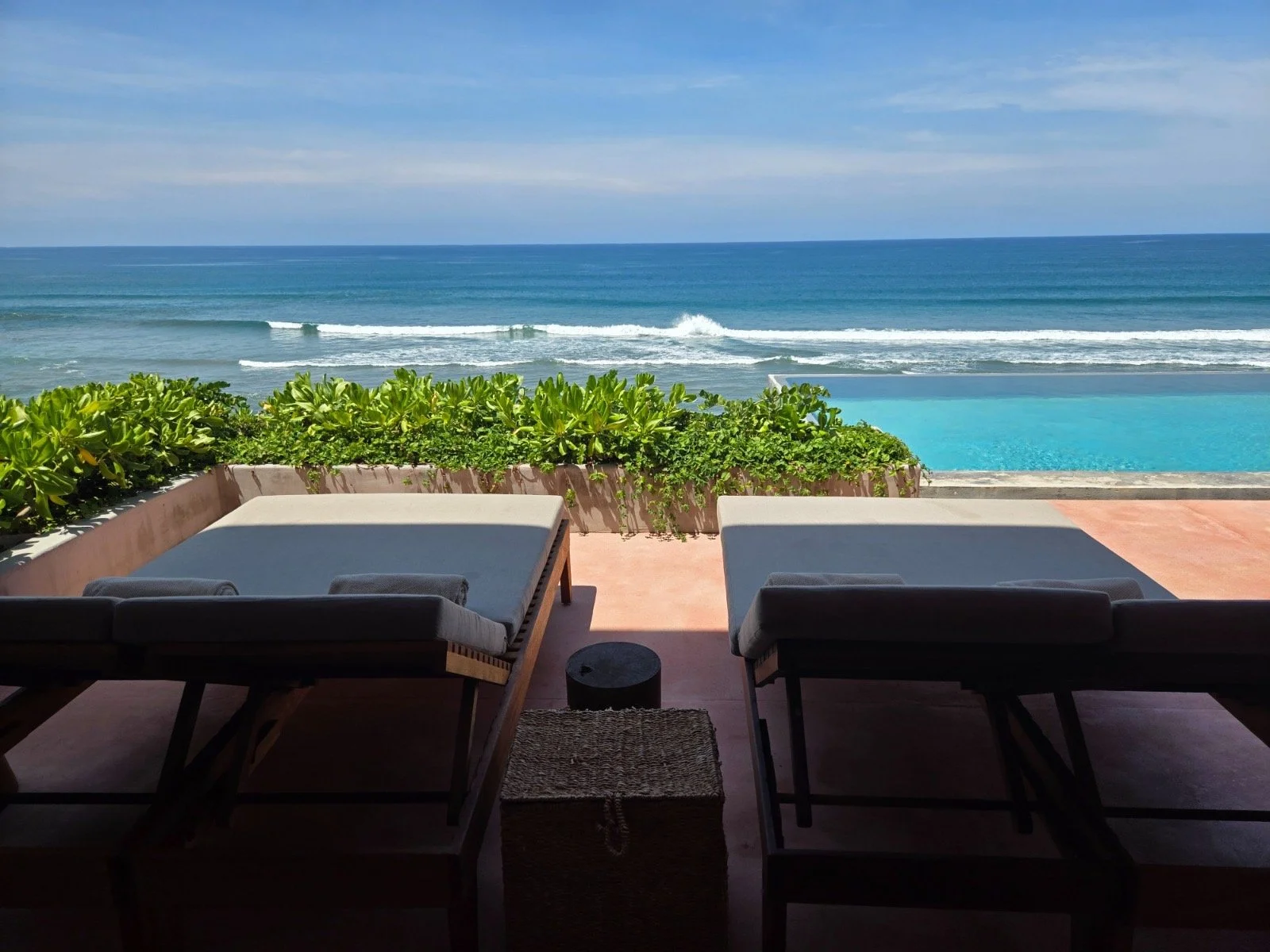 View from a patio overlooking a swimming pool, greenery, and the ocean with waves, under a partly cloudy sky.