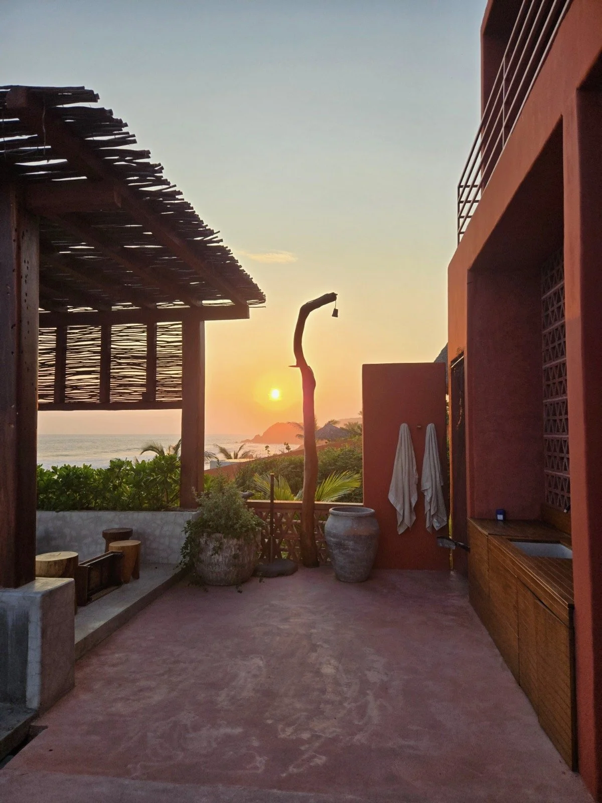 Sunset view from a terrace with ocean in the background, palm trees, rustic wooden pergola, potted plants, towels hanging on a wall, and a sculptural wooden stick with a small bell hanging from it.