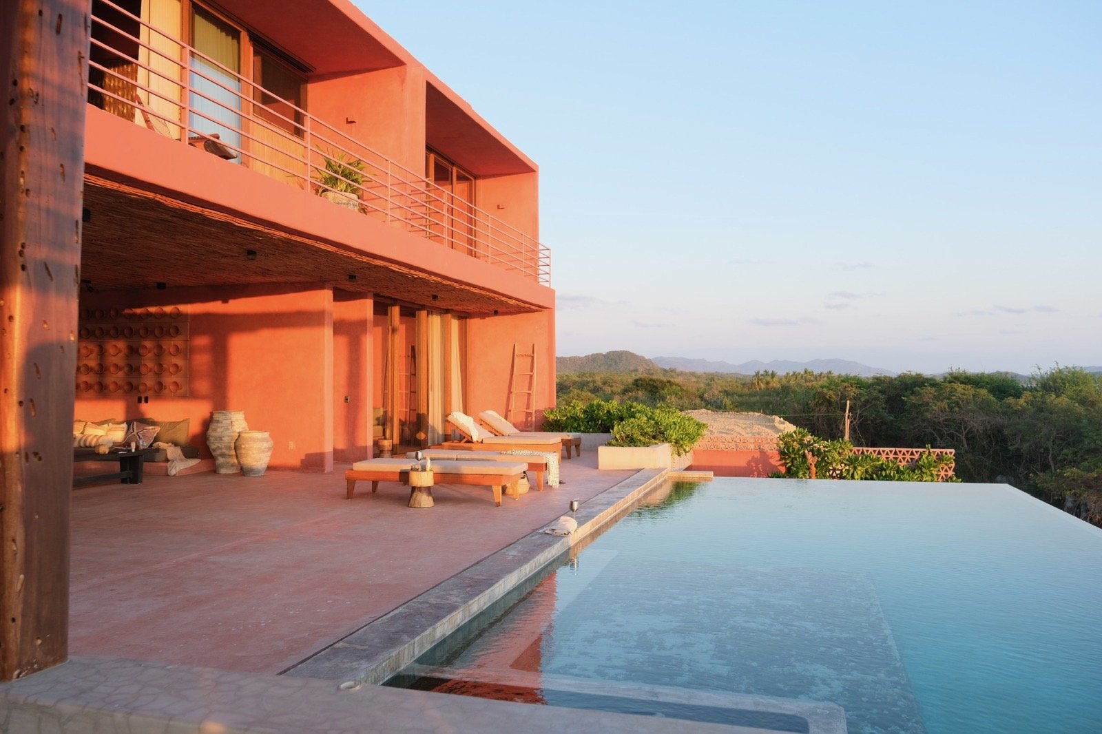 A modern pink stucco house with a swimming pool in the foreground, surrounded by lush greenery and distant mountains under a clear sky.