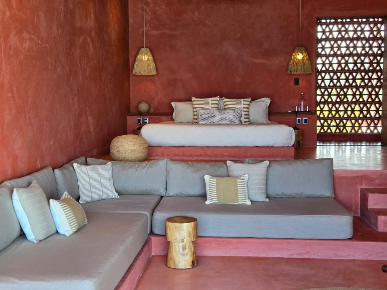 Living room with red textured walls, a platform bed with pillows, gray cushions and a small wooden stool, natural woven pendant lights, and a decorative wooden screen with geometric cutouts.