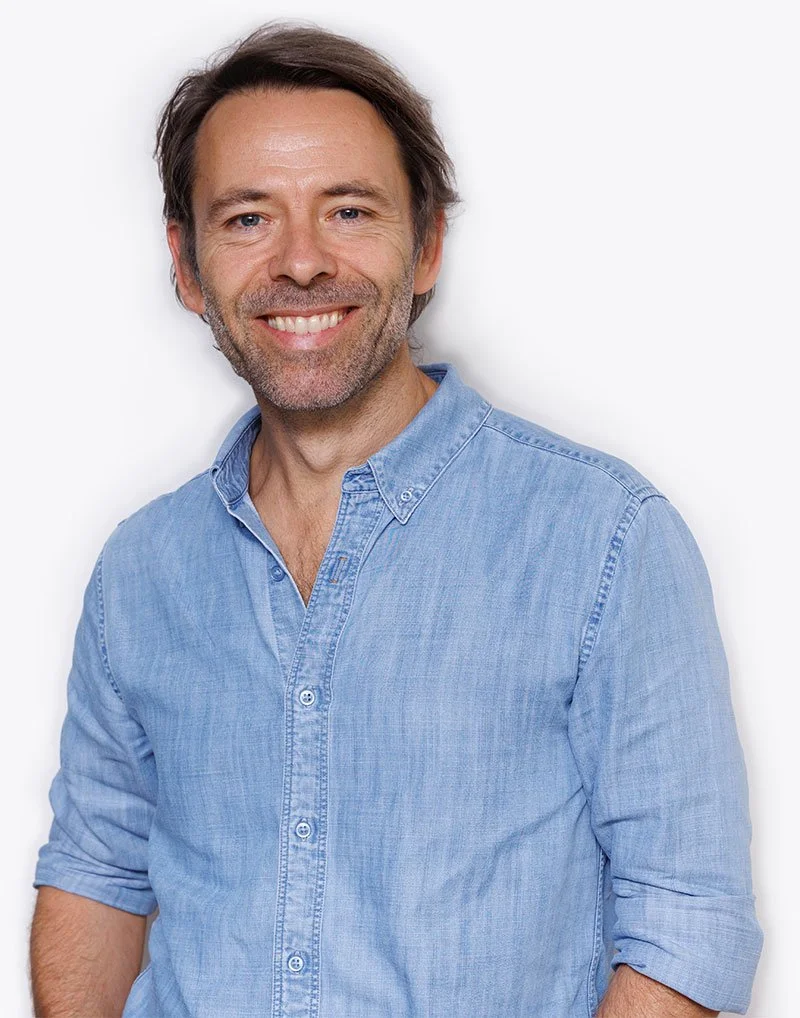 A man in a light blue denim shirt smiling against a plain white background.