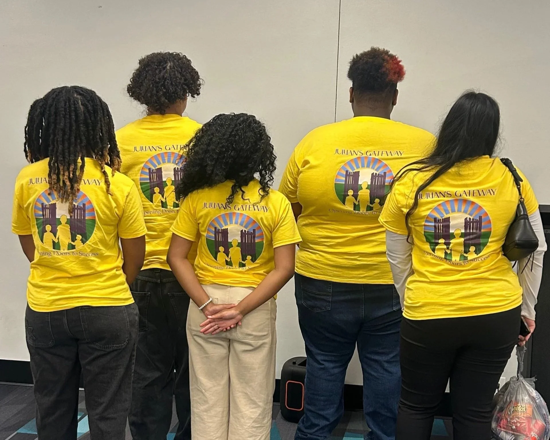 Group of five people standing with backs to the camera, wearing yellow t-shirts with a logo and text that reads "Julian's Gateway" and "Opening Doors to Success." They are in a room with a plain white wall.