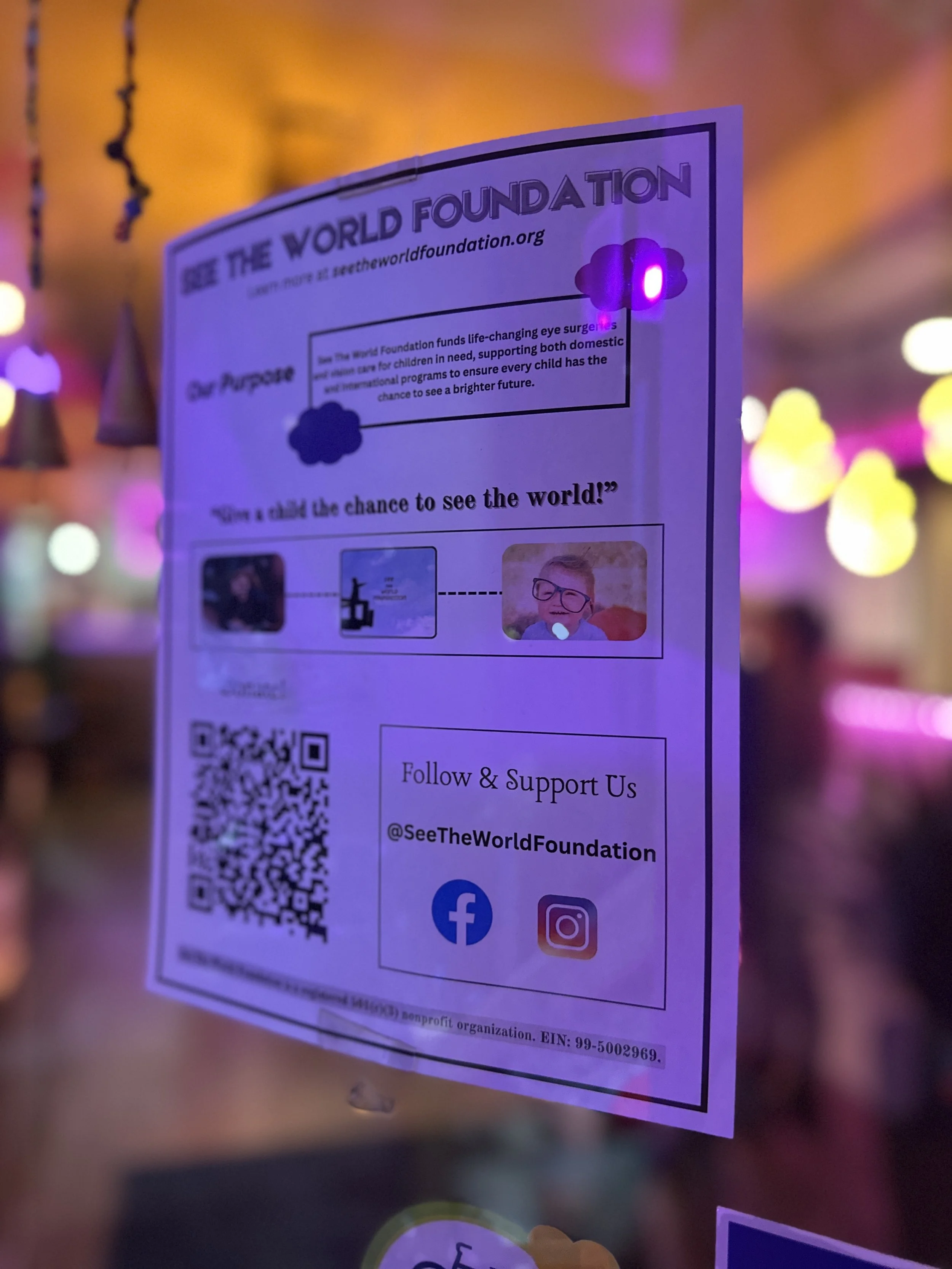 A flyer for the See The World Foundation, promoting eye surgery for children in need, with a QR code and social media icons for Facebook and Instagram, displayed on a glass surface with colorful blurred lights in the background.