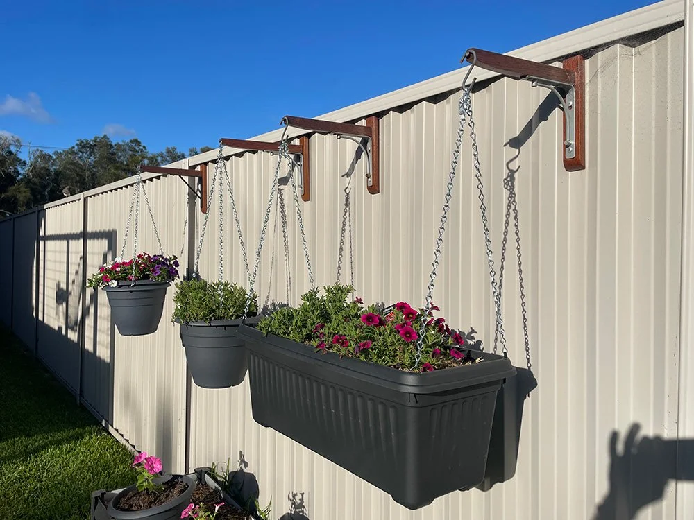 Three hanging flower pots with pink and purple flowers mounted on a white fence under a blue sky.