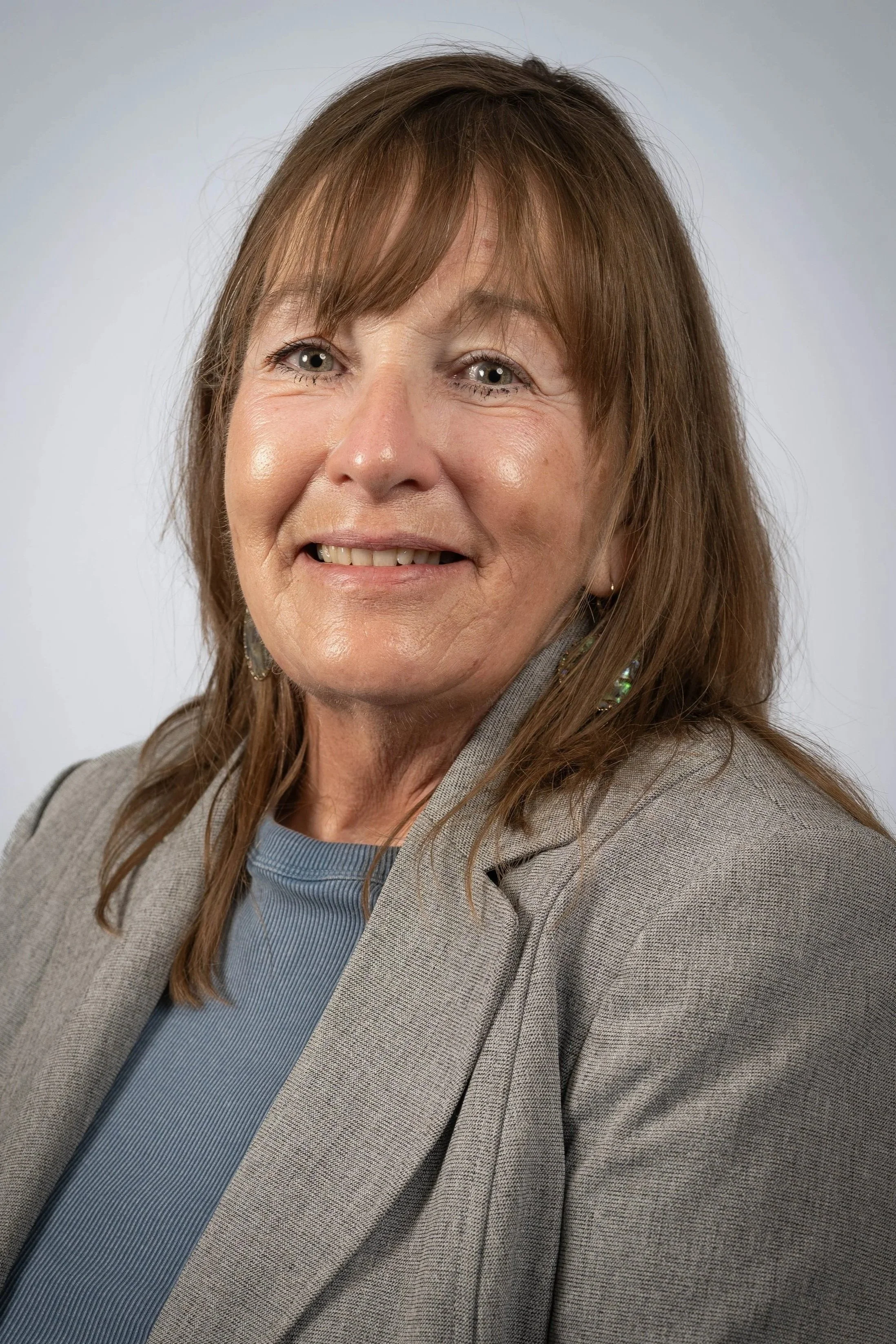 A smiling woman with mid length brown hair and bangs, wearing a neutral color blazer