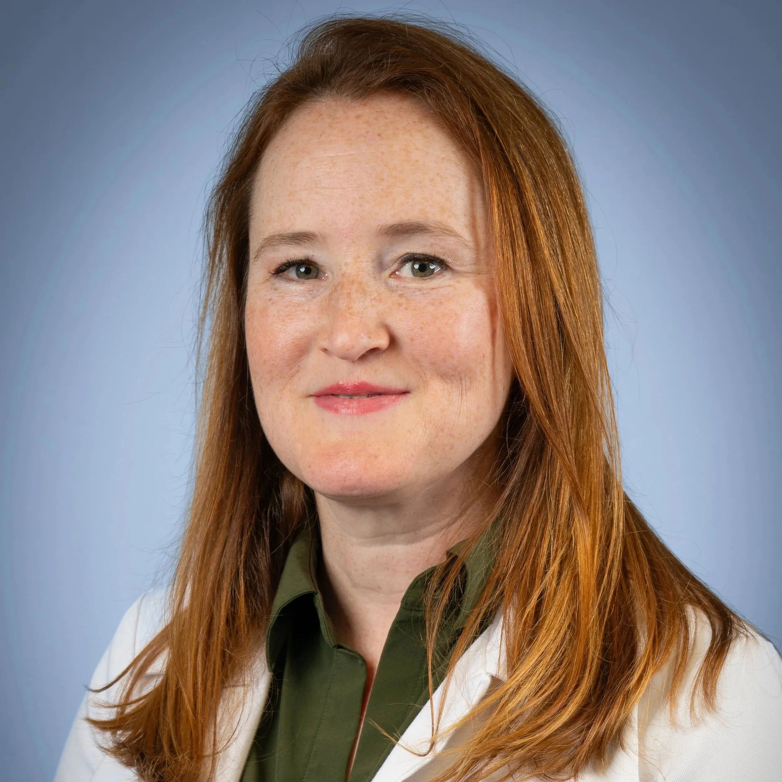 A headshot of a smiling woman shoulder-length, reddish hair, wearing a green top and white provider coat, with a blue neutral background. 