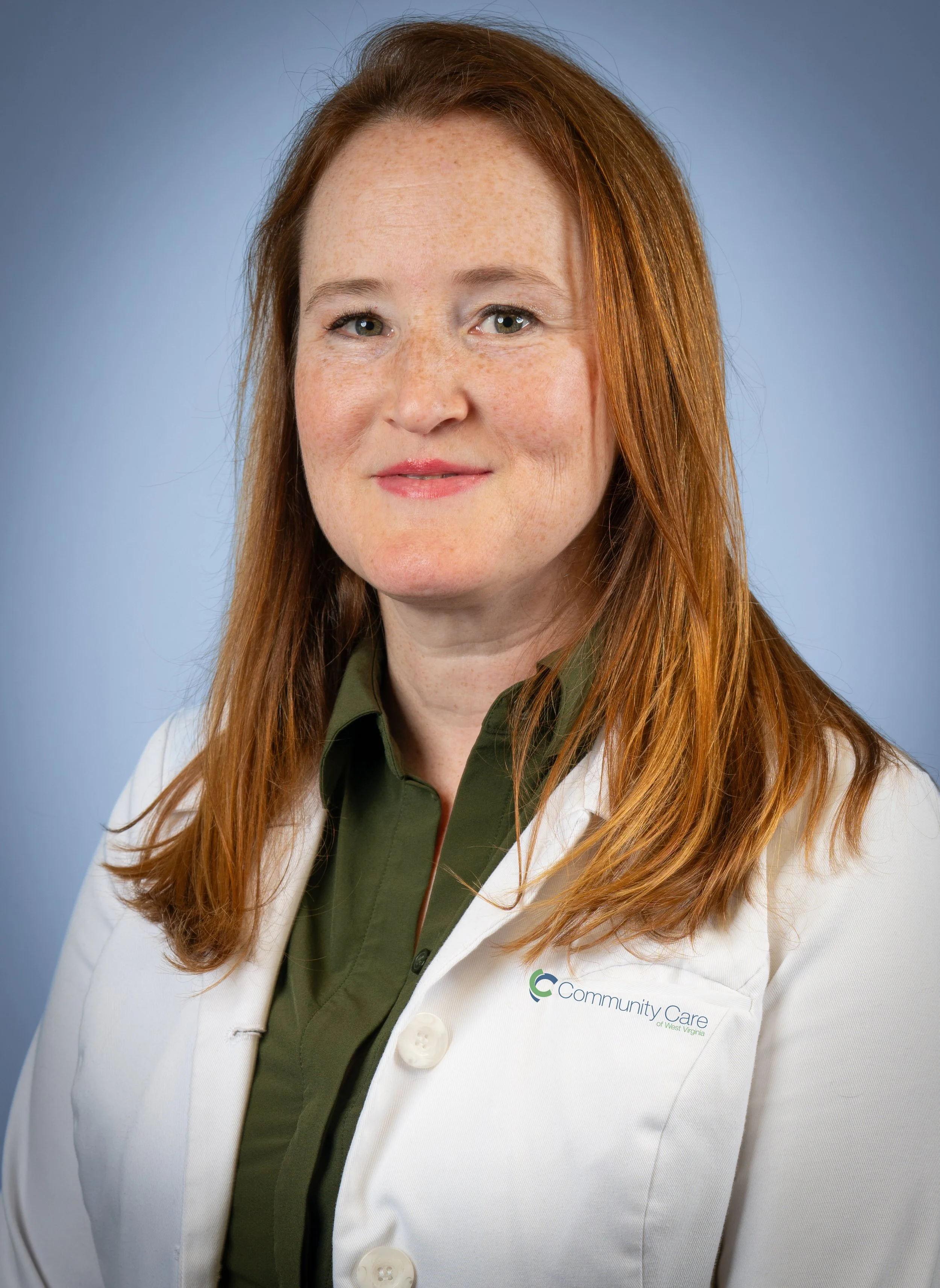 A headshot of a smiling woman shoulder-length, reddish hair, wearing a green top and white provider coat, with a blue neutral background.