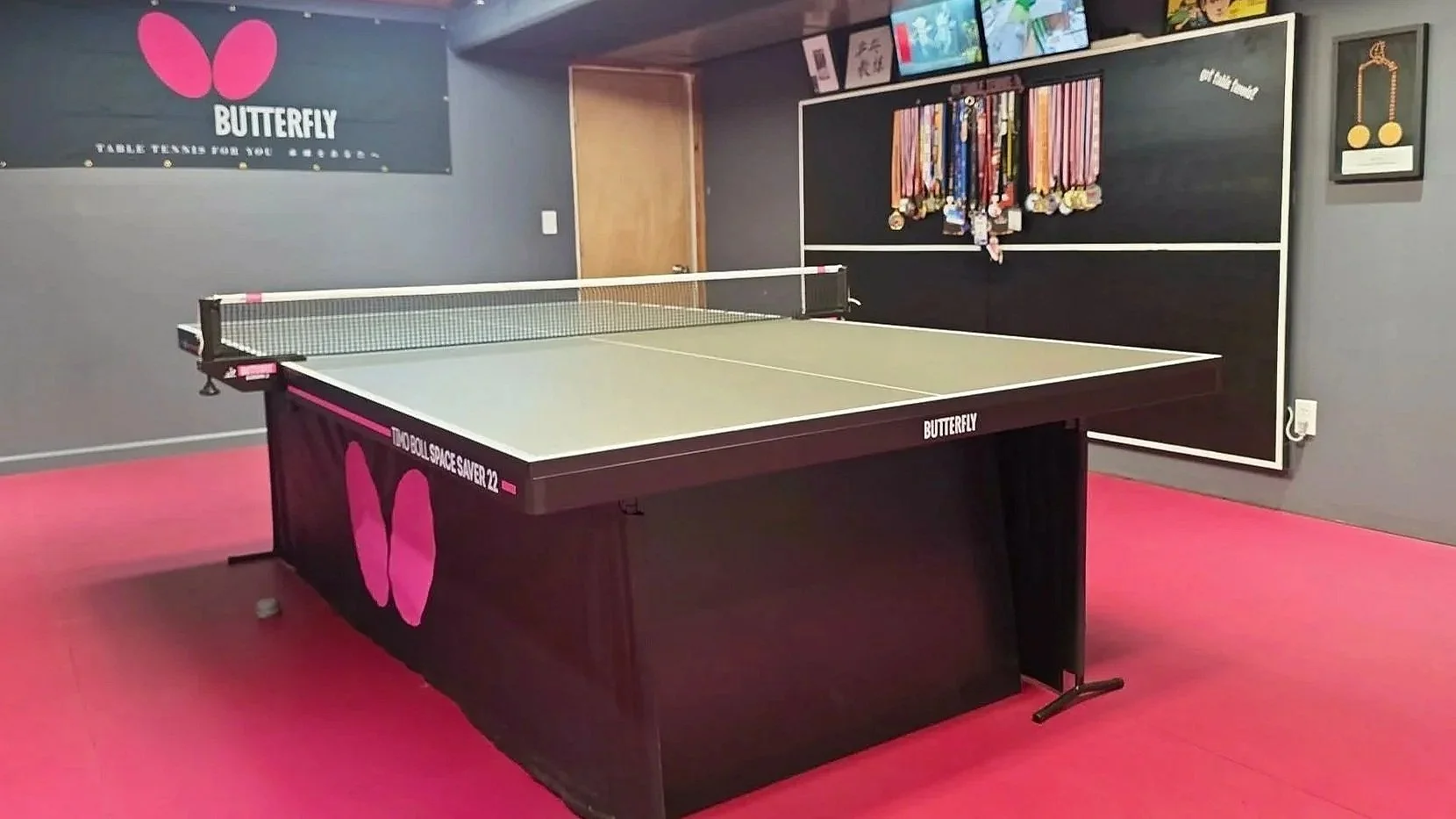 Indoor room with a butterfly brand table tennis table on pink flooring, medals hanging on a black wall panel, and a media shelf with several television screens above the medals.
