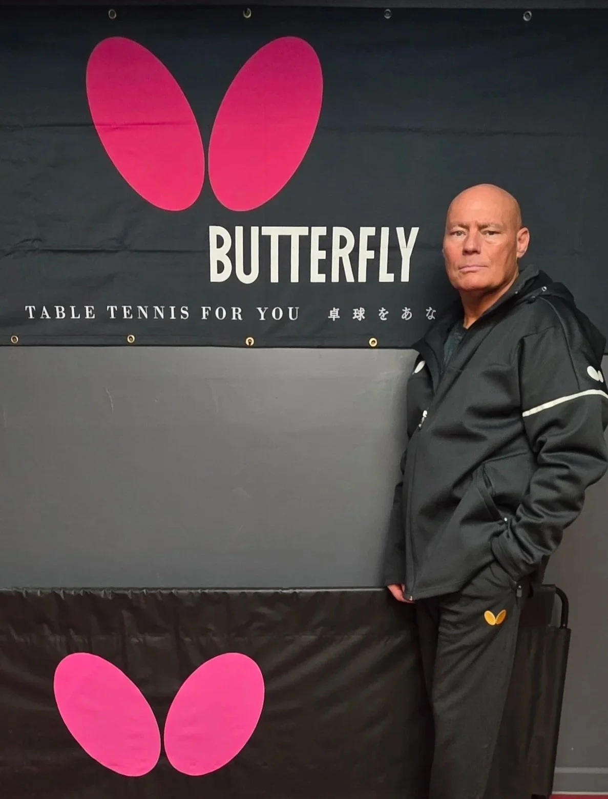 Coach Meinhard standing next to a black table tennis banner with a pink butterfly logo and the words "Butterfly Table Tennis for You" in English and Japanese.