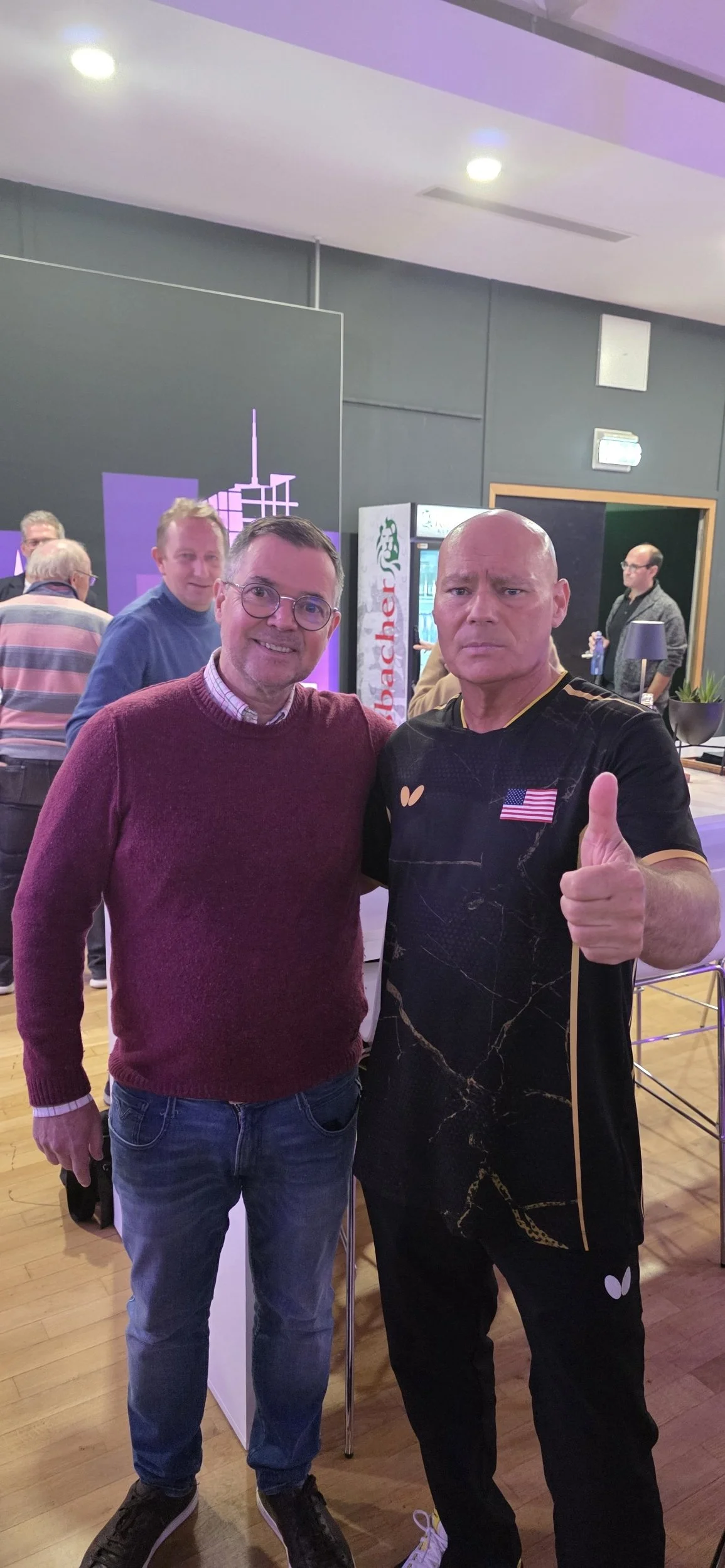 Coach Meinhard with Speedy posing for a photo at an indoor event, with a background featuring a purple cityscape graphic, vending machines, and other attendees.